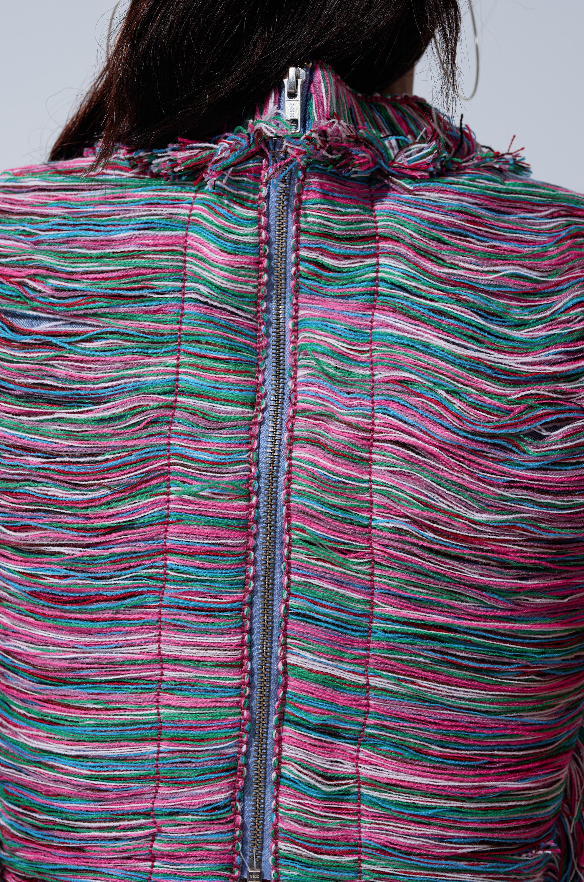 Close-up of the back of the SHES GONE, DANCE ON GIRL SHREDDED DENIM TOP with textured multicolored horizontal threads in pink, green, blue, and purple, a visible central zipper, and partially visible dark hair at the top.