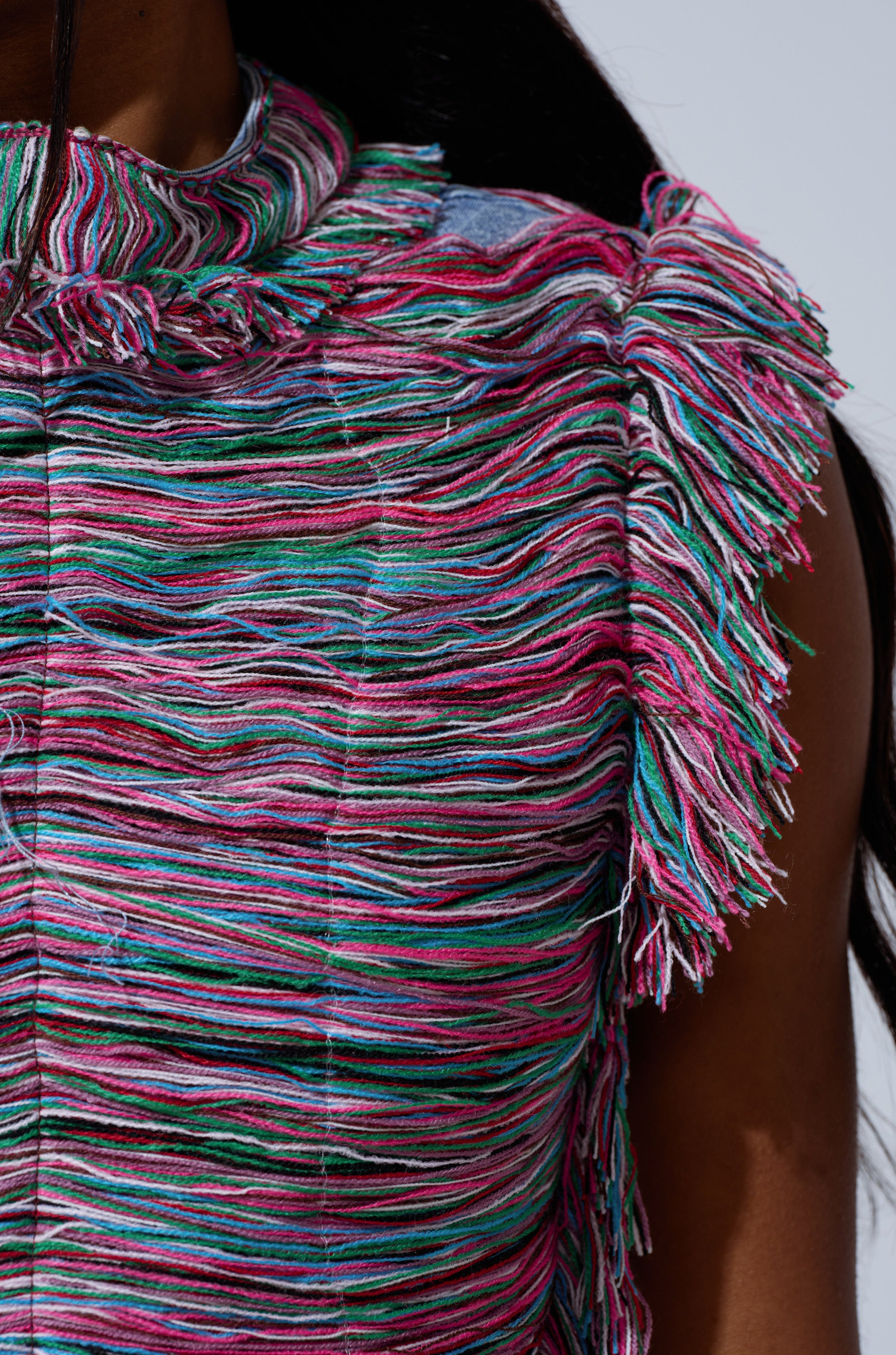 Close-up of a person wearing the SHES GONE, DANCE ON GIRL SHREDDED DENIM TOP, featuring a colorful, textured cropped design with layered strands of pink, blue, green, and white thread and multicolored yarn fringe on the shoulder and collar.