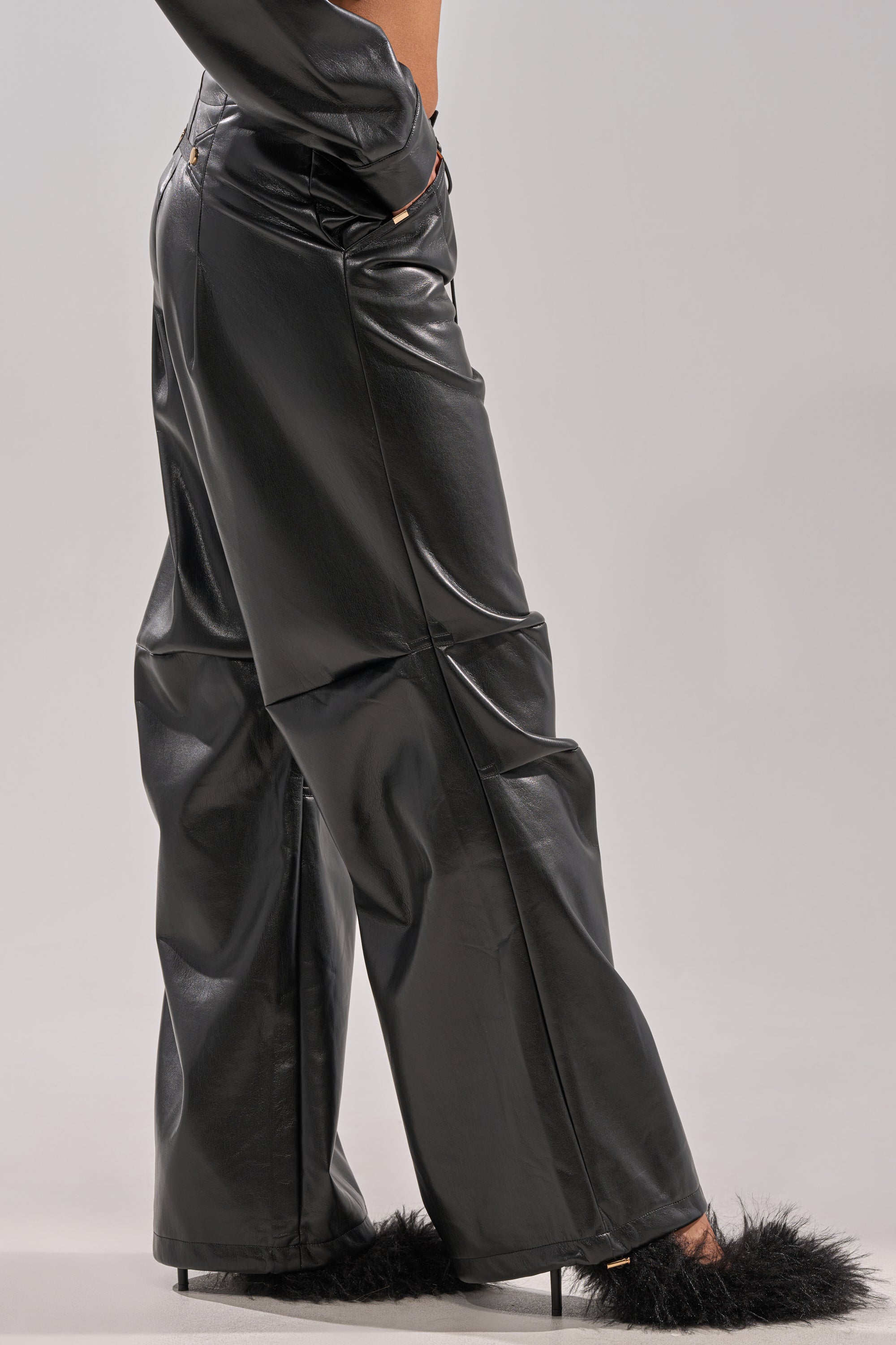 A person wears the GET WITH IT FAUX LEATHER TROUSER in shiny black and black fuzzy high-heeled shoes, standing against a plain light gray background.
