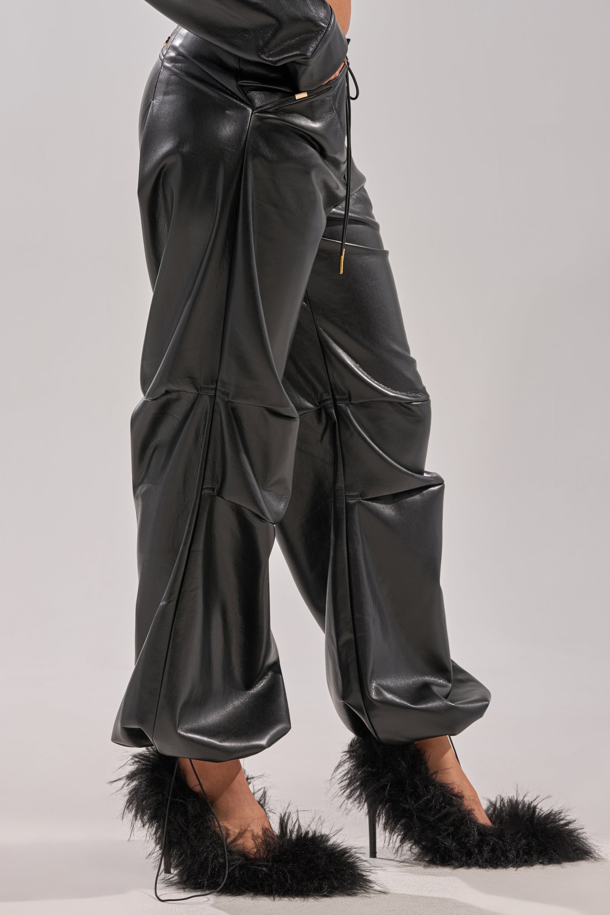 A person wearing GET WITH IT FAUX LEATHER TROUSER in shiny black and high-heeled shoes with black fluffy details, standing against a light background.