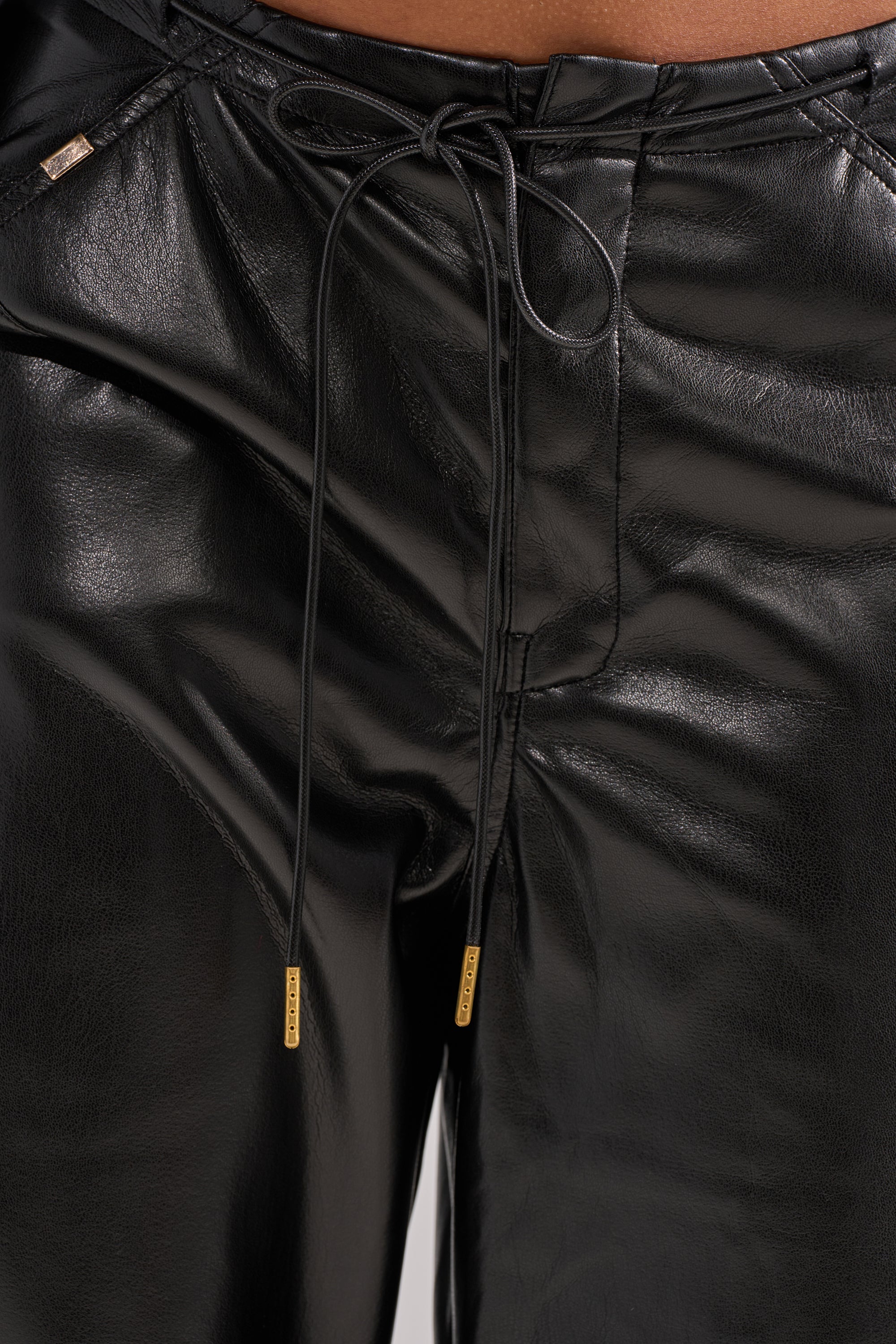 Close-up of a person wearing the GET WITH IT FAUX LEATHER TROUSER, featuring shiny black faux leather, high-rise fit, drawstring waist with metallic tips, smooth texture, and wide-leg style.