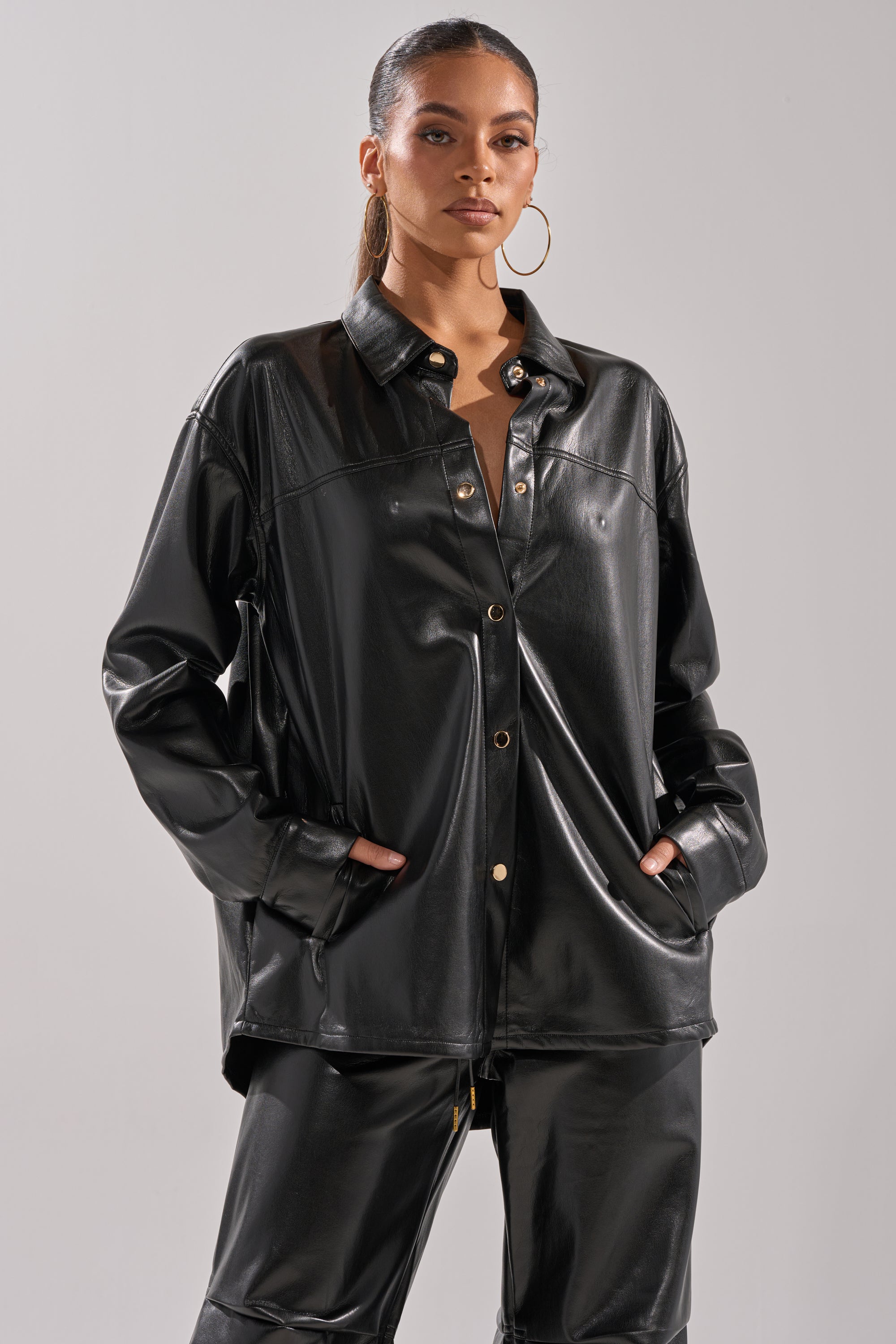 A woman wears the GET WITH IT FAUX LEATHER BUTTON DOWN with gold snap-buttons and matching black faux leather pants, hands in her pockets, posing confidently against a plain light gray background.