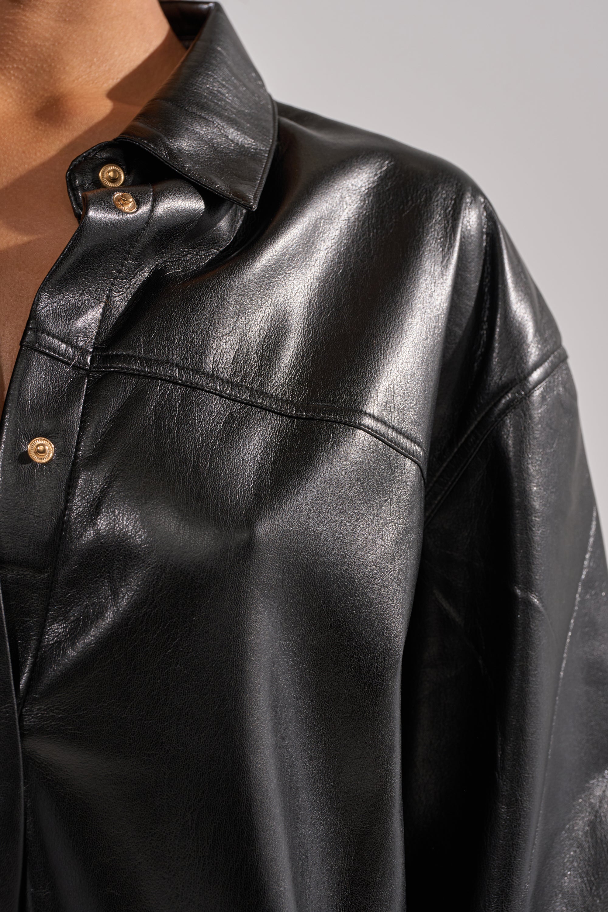 Close-up of a person wearing the GET WITH IT FAUX LEATHER BUTTON DOWN in shiny black, featuring a collared neckline and gold buttons. The image captures the material's glossy texture, with only the upper body and part of the face visible.