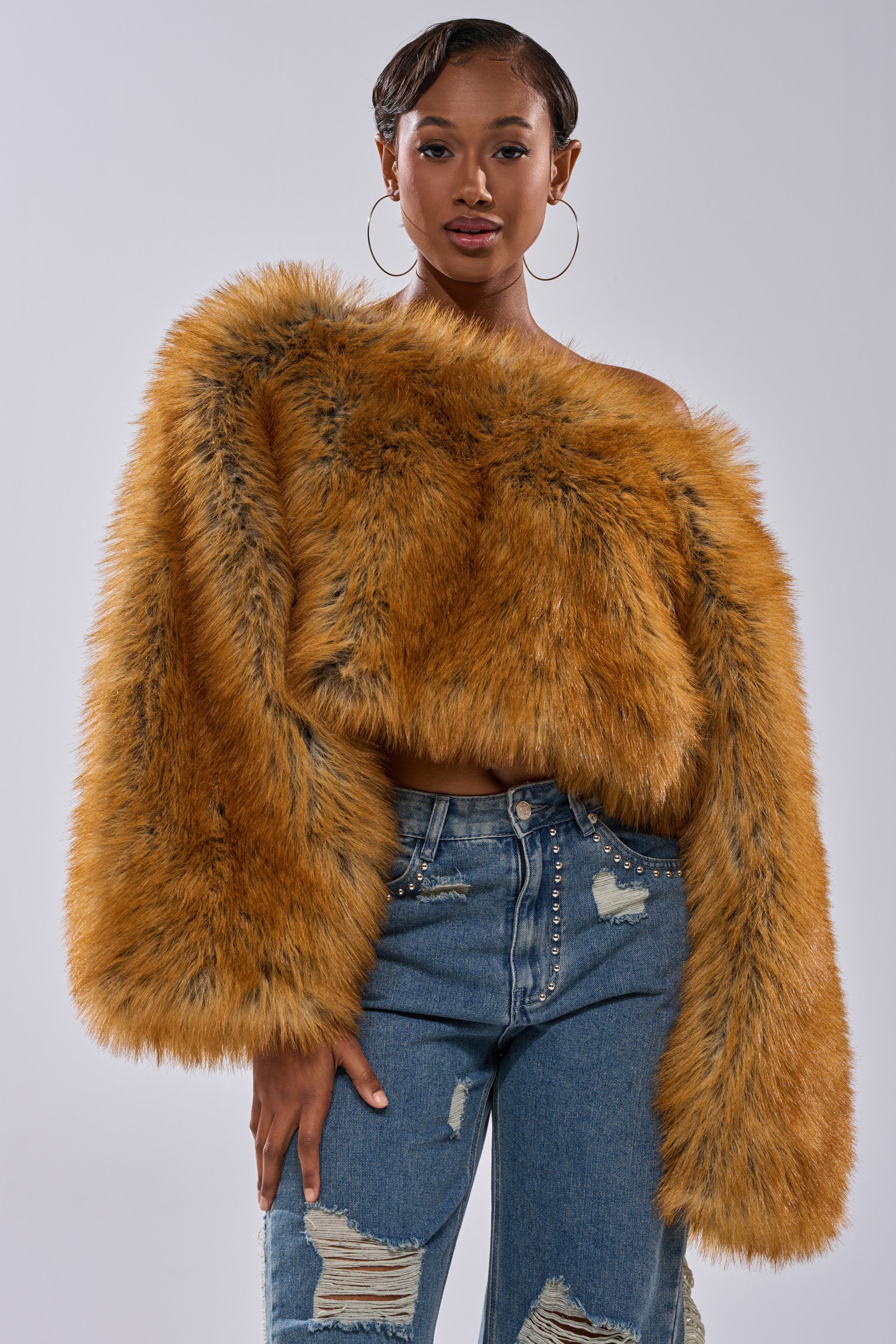 A woman wears large hoop earrings, ripped blue jeans, and the SNOW DAY FAUX FUR SWEATER off-shoulder, confidently posing against a plain light gray background.