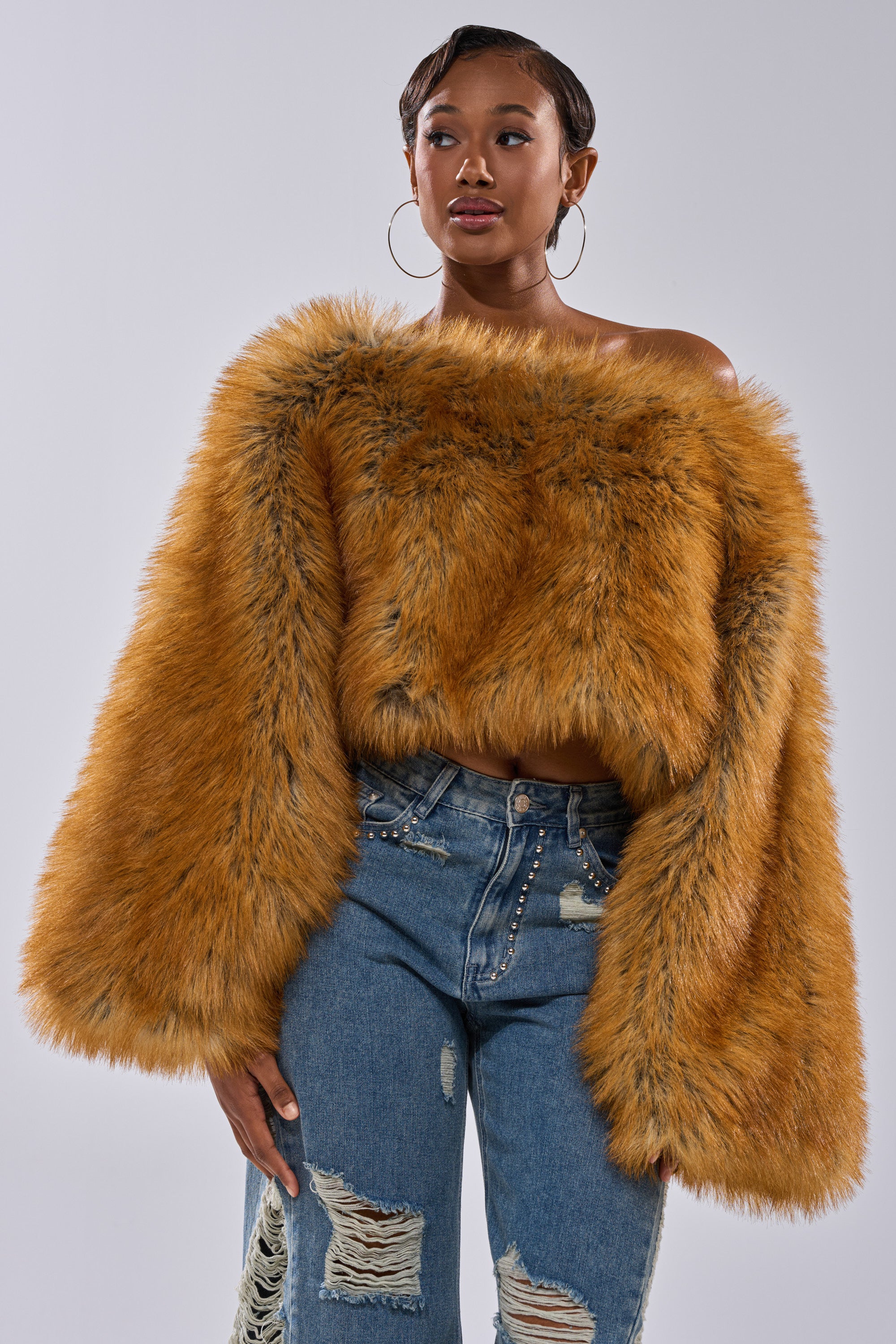 A woman models winter-chic style in the SNOW DAY FAUX FUR SWEATER in orange, paired with blue distressed jeans embellished with pearls, against a plain light gray background.