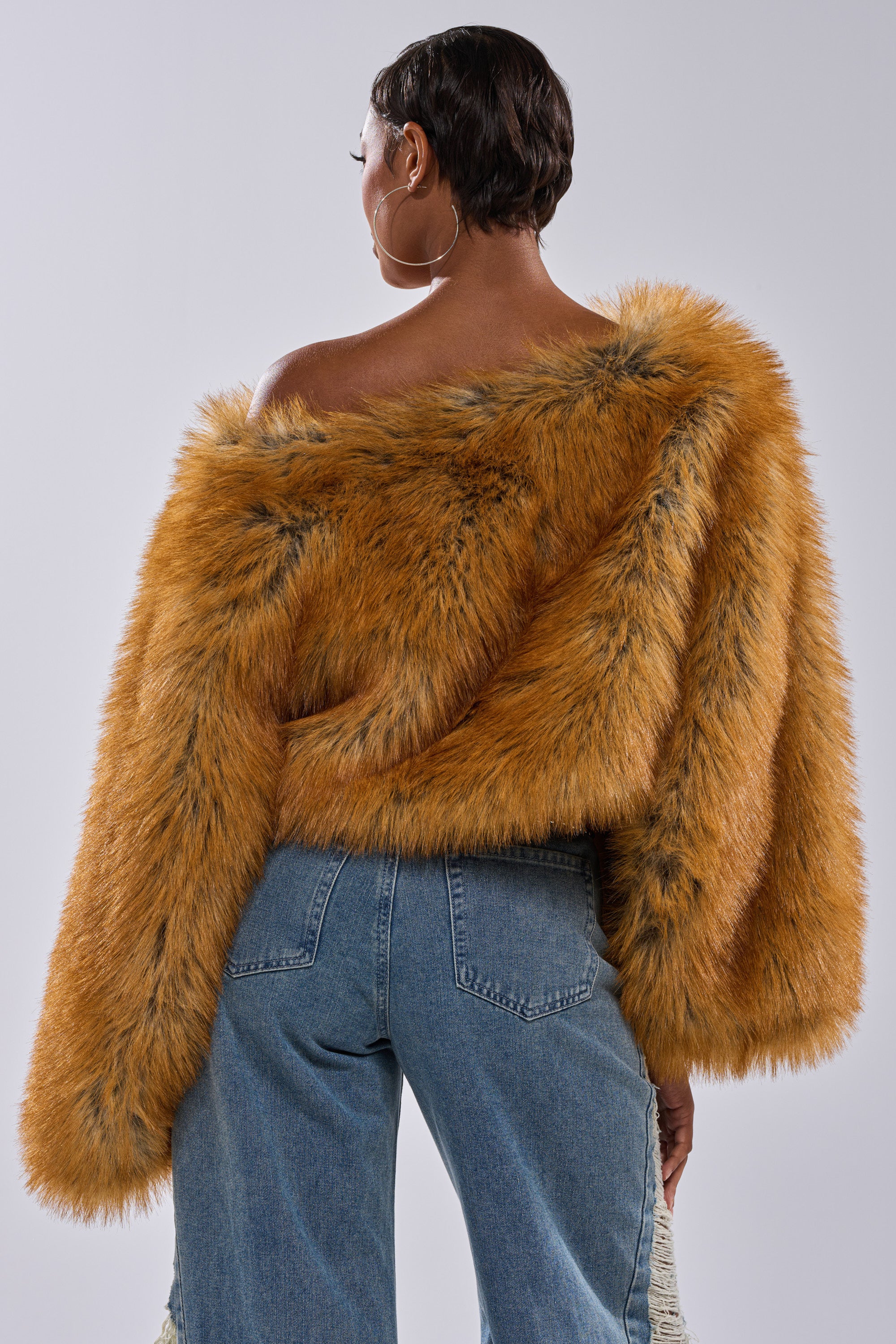 A person with short hair is seen from behind wearing large hoop earrings, high-waisted blue jeans, and the SNOW DAY FAUX FUR SWEATER in orange-brown, styled off the shoulder, against a plain light background.