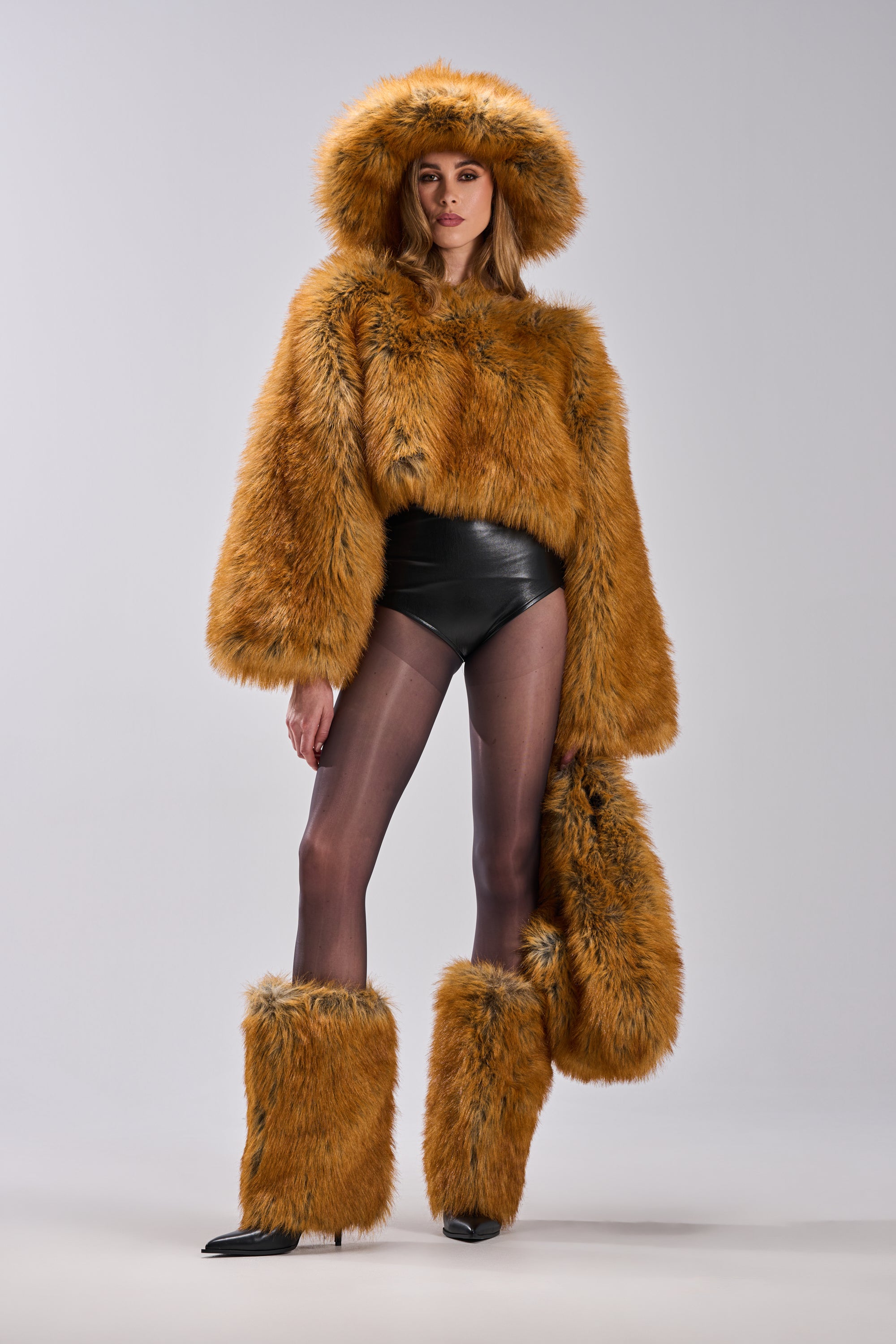 A person stands against a light-gray background wearing the SNOW DAY FAUX FUR SWEATER, matching faux fur boots and mittens, black leather shorts, and sheer black tights.