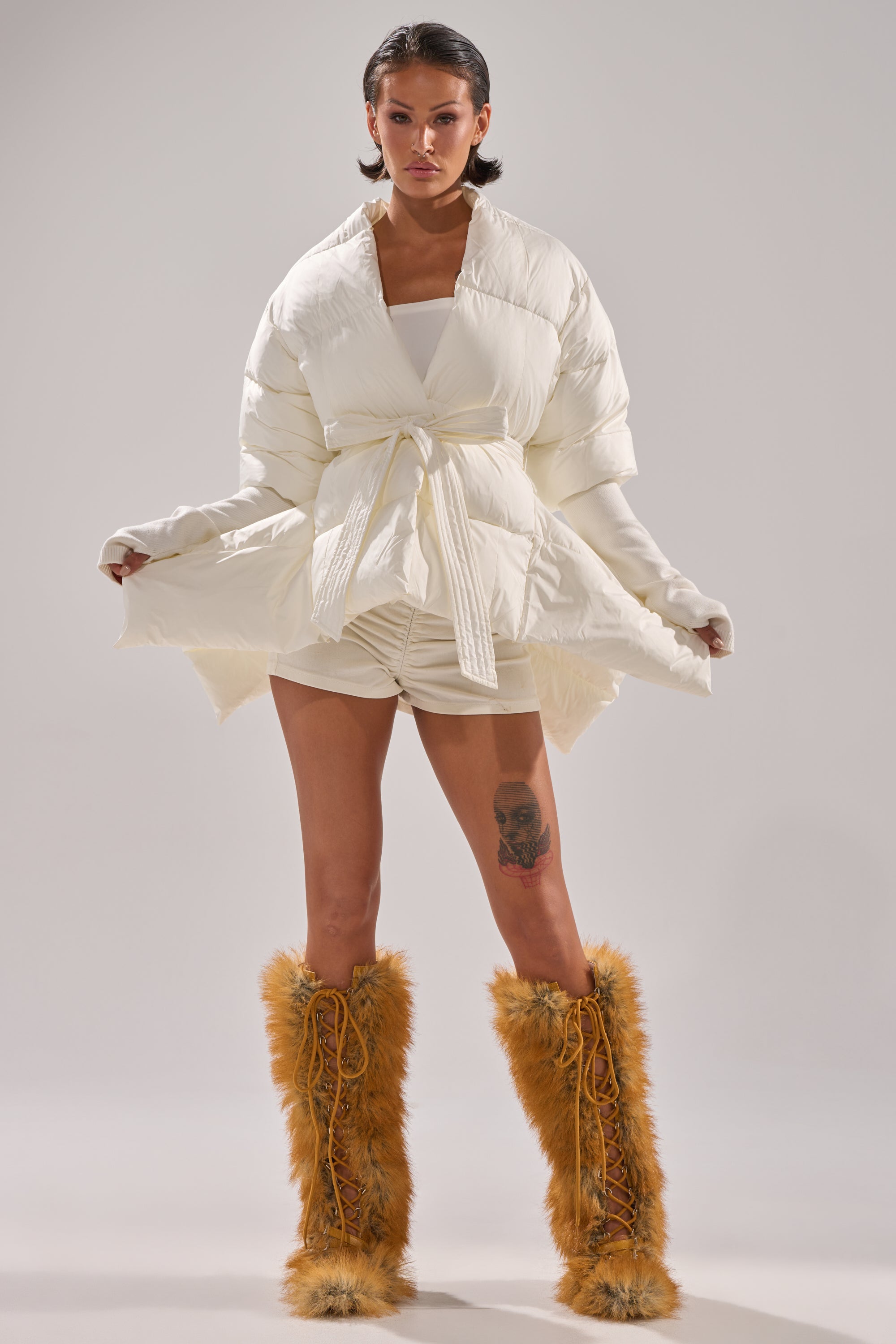 A woman confidently poses in the TALK TO ME NICE FUR TRIM PUFFER, styled with white ruched shorts and tall tan furry boots, against a plain light background.