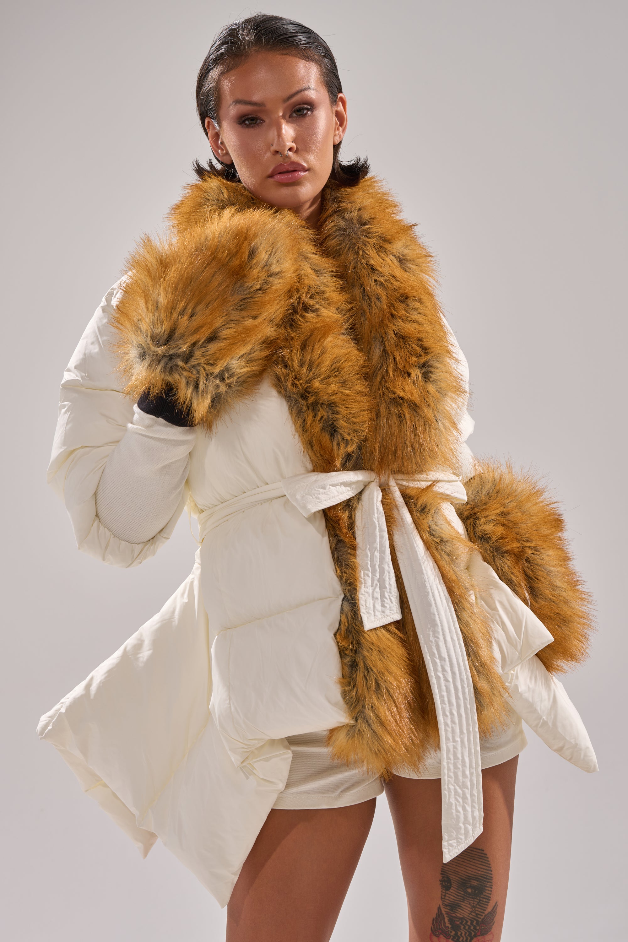 A person models the TALK TO ME NICE FUR TRIM PUFFER, a white belted coat with brown faux fur trim and cuffs, posing confidently against a plain light background.