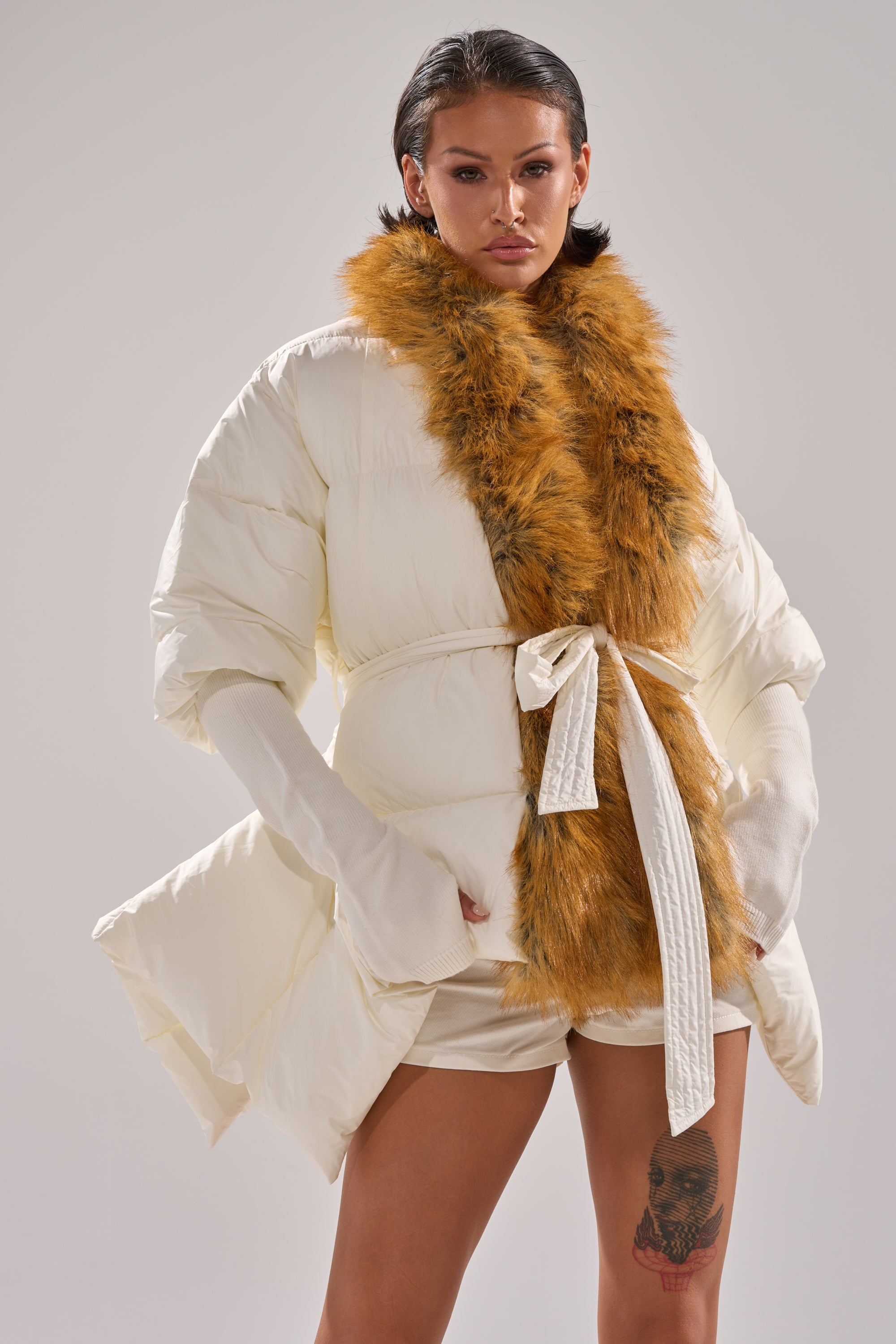 A person with short dark hair and a thigh tattoo models the TALK TO ME NICE FUR TRIM PUFFER, featuring white fur trim and a brown faux fur collar, belted at the waist over white shorts, against a plain light background.
