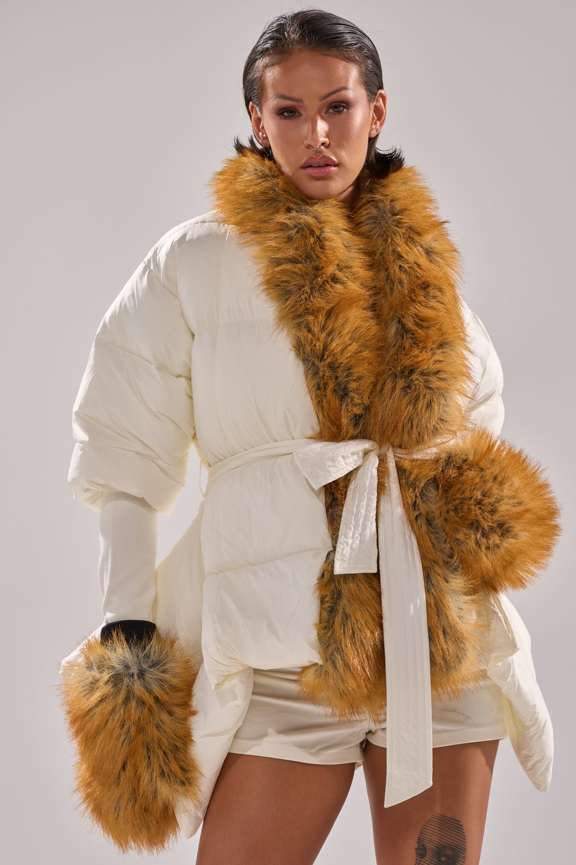 A woman models the TALK TO ME NICE FUR TRIM PUFFER, featuring a white puffer design with faux fur trim, a wide belt, and fur-cuffed sleeves. She pairs it with beige shorts and slicked-back hair against a neutral background.