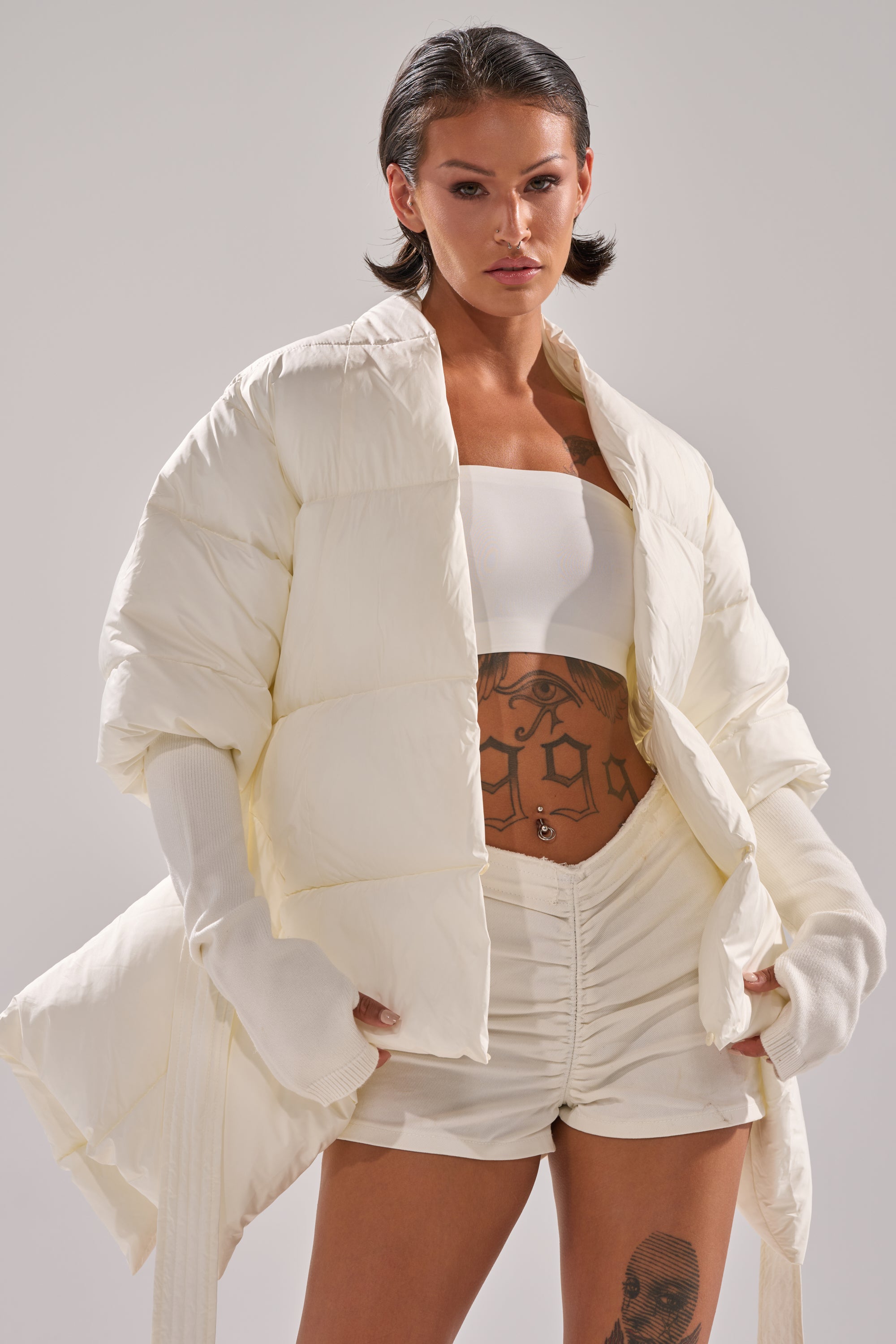 Wearing the TALK TO ME NICE FUR TRIM PUFFER over a white bandeau top and ruched white shorts, a tattooed model poses confidently against a plain background.