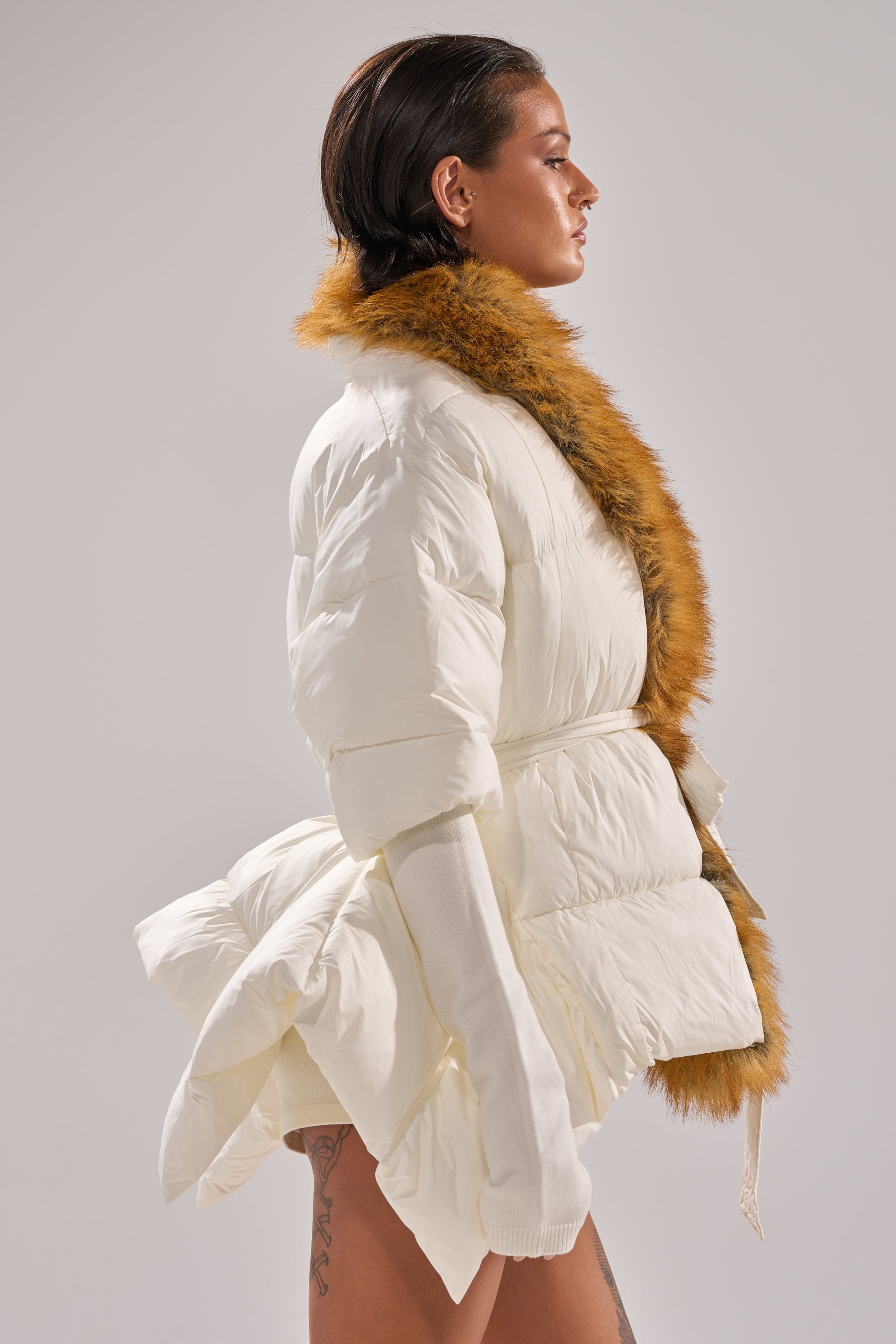 A woman with slicked-back dark hair and a thigh tattoo models the TALK TO ME NICE FUR TRIM PUFFER—a white puffer jacket with a brown faux fur collar and long sleeves—standing against a plain light gray background.
