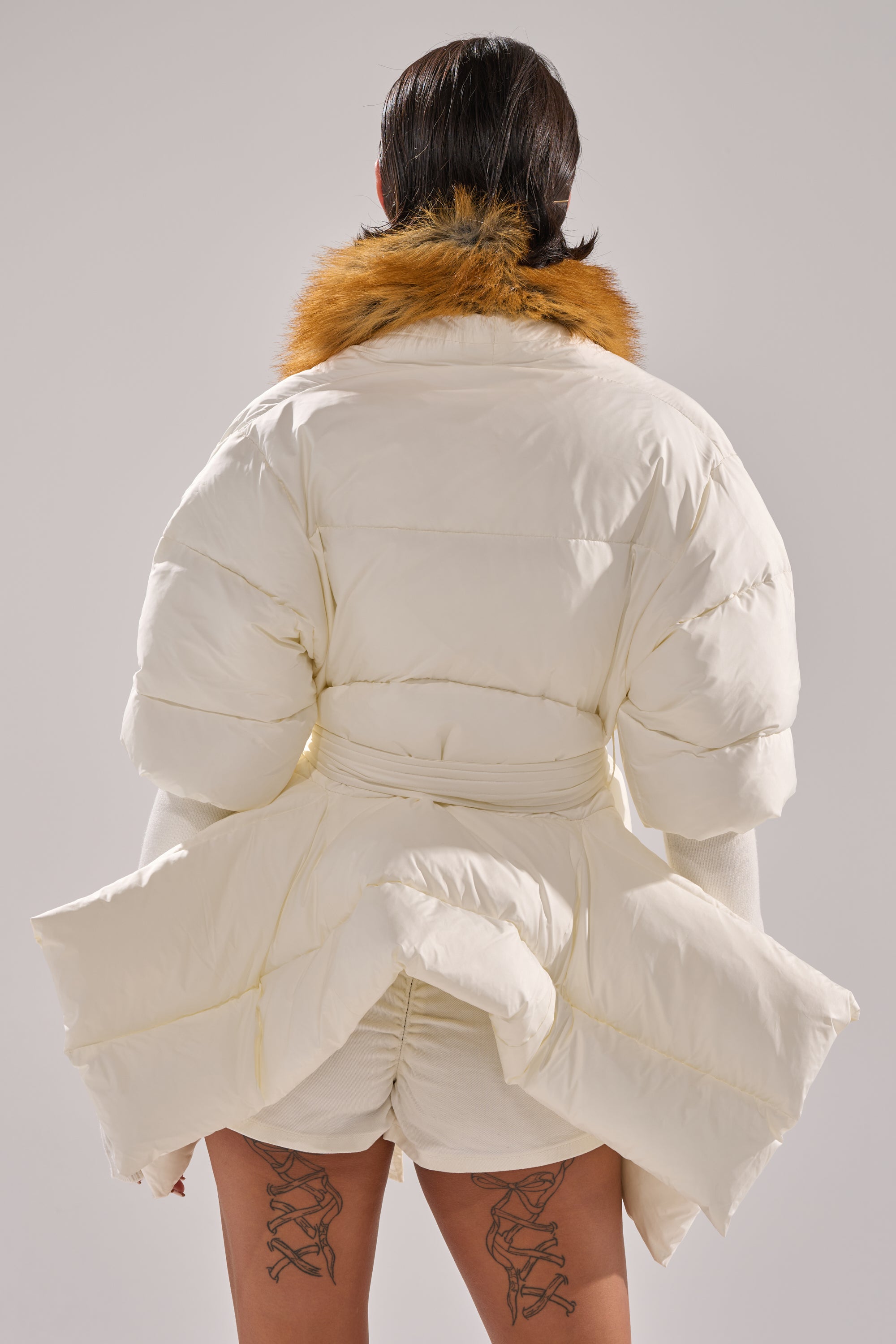 A person is seen from behind wearing the TALK TO ME NICE FUR TRIM PUFFER—a white puffer jacket with a brown faux fur collar and bold sculptural design—plus crossed ribbon tattoos on the backs of their thighs.