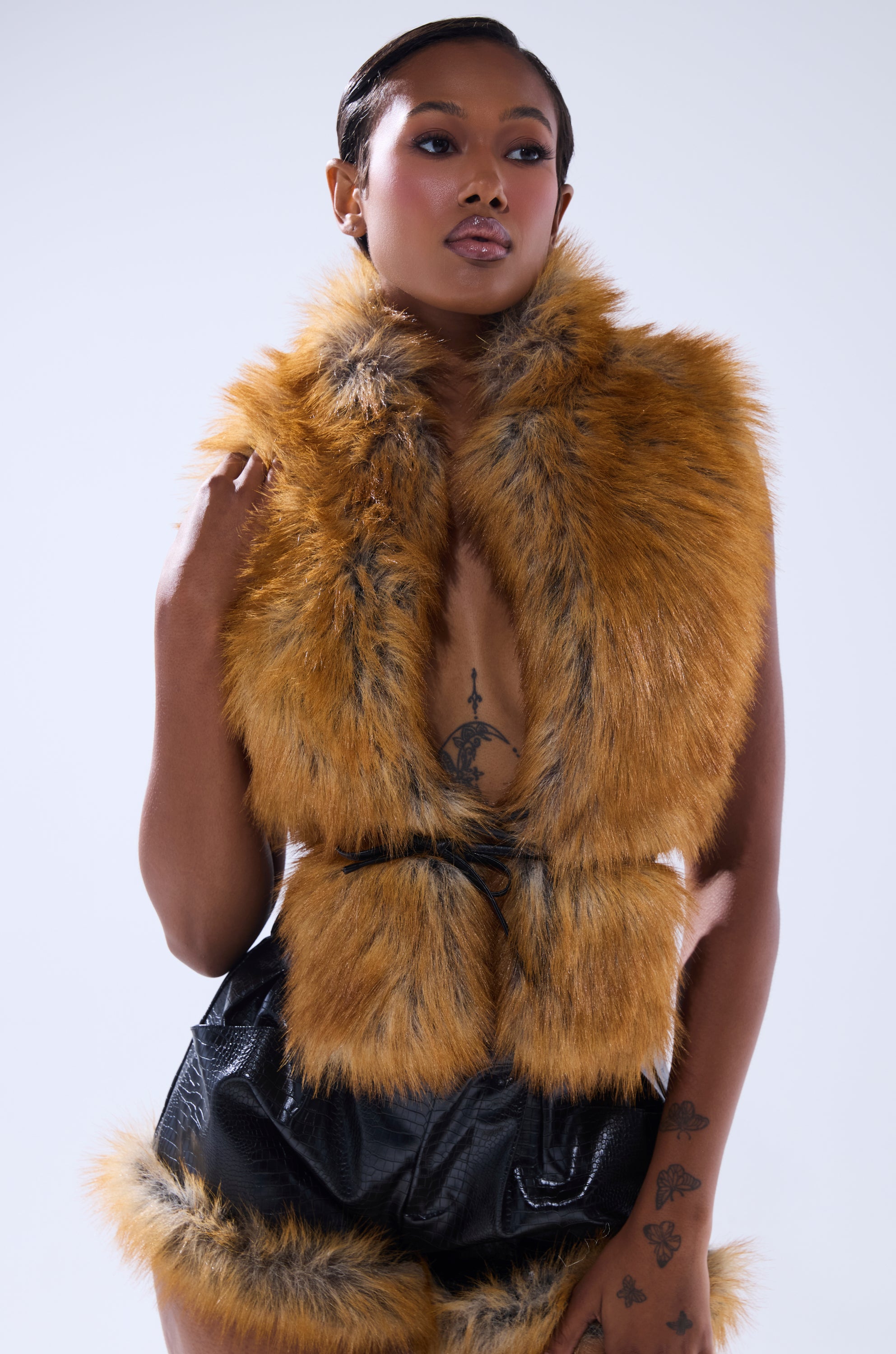 A person poses confidently in the DONT TOUCH OR LOOK FUR BLOUSE, featuring a leather tie closure, paired with high-waisted black shorts. They show tattoos on their chest and hand, with short styled hair against a neutral background.