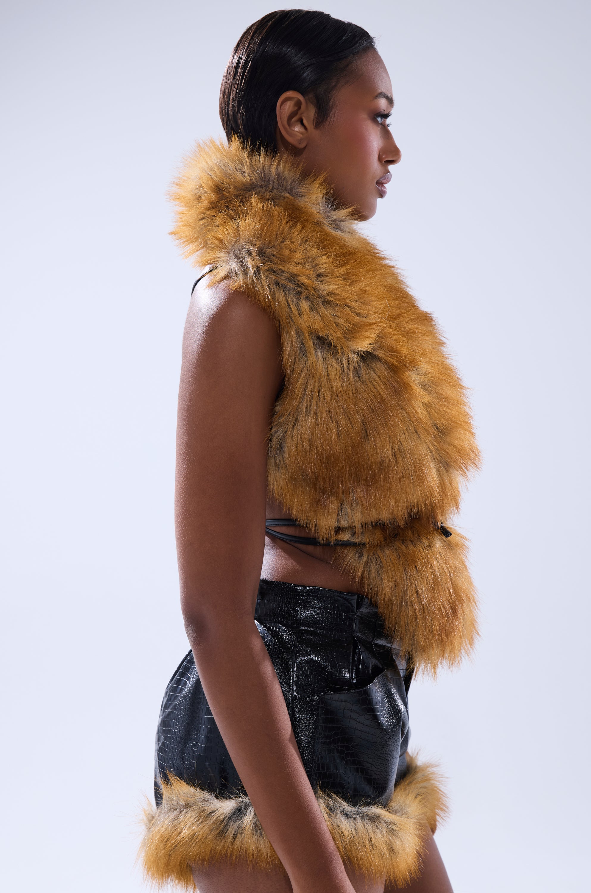 A woman in profile models the DONT TOUCH OR LOOK FUR BLOUSE, a golden brown faux fur vest with a leather tie closure, paired with matching fur-trimmed black shorts against a plain light background.