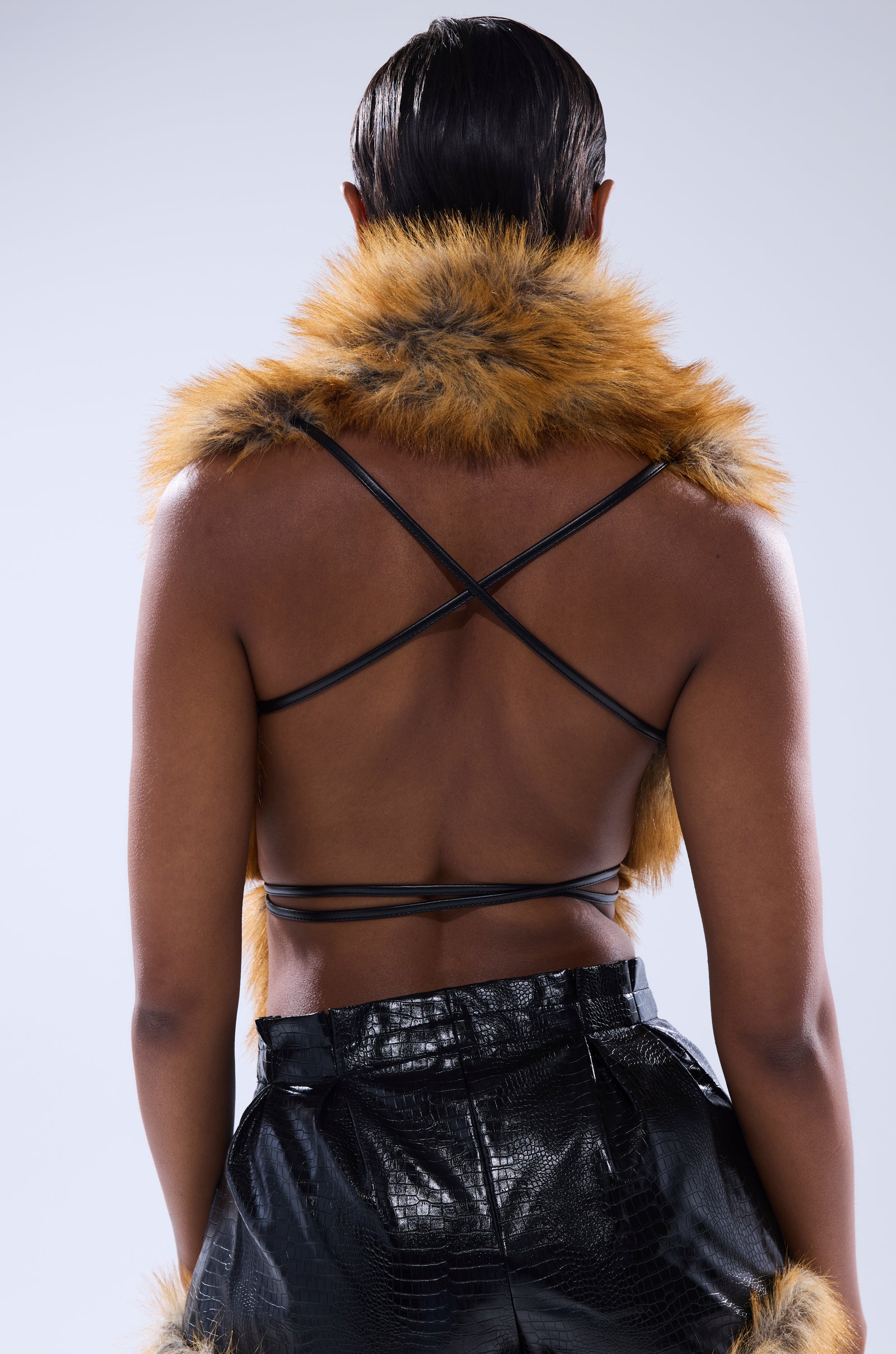 A person with short, slicked-back hair is seen from behind wearing the DONT TOUCH OR LOOK FUR BLOUSE, which features straps and brown shoulder accents, paired with shiny black high-waisted pants.