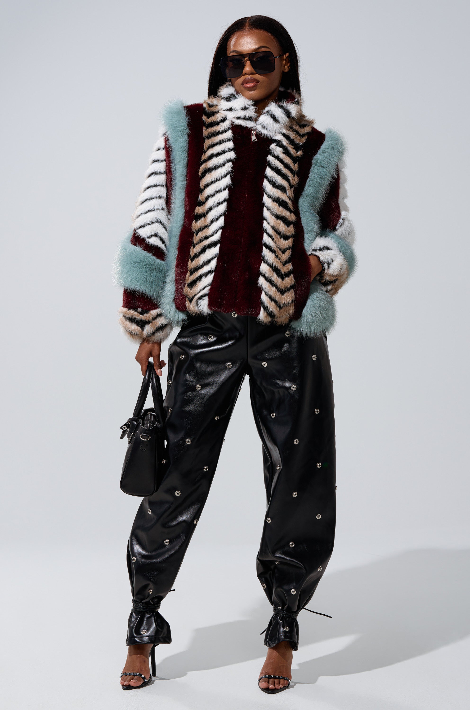 A stylish woman stands confidently in the HAVE IT ALL PANELED FUR COAT, black leather pants with metallic accents, heeled sandals, sunglasses, and a small black handbag against a plain background.