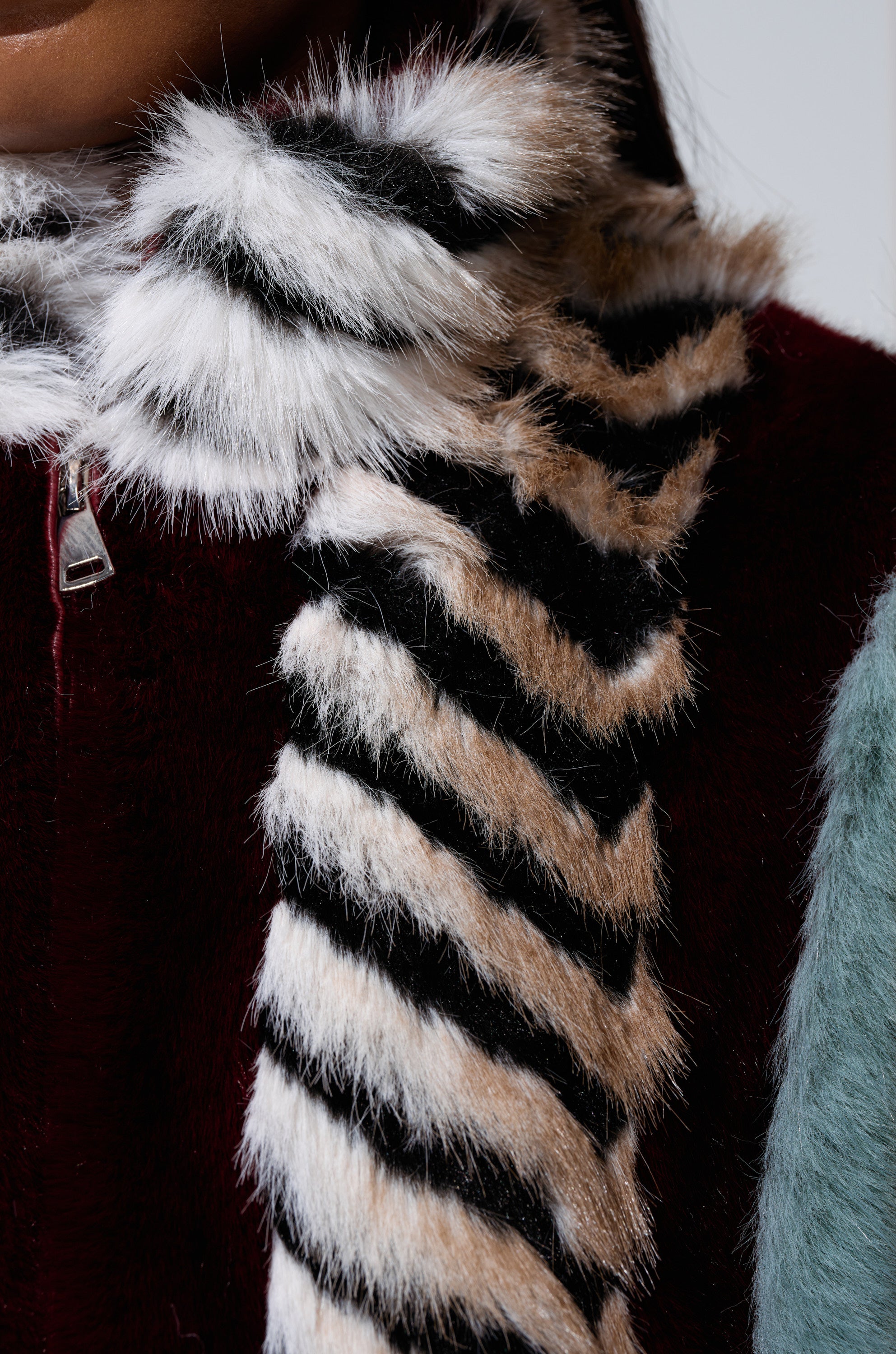 Close-up of a person in the dark red HAVE IT ALL PANELED FUR COAT with a zipper, styled with a striped black, white, and beige faux fur scarf. Teal fuzzy fabric on the right side adds contrasting texture.