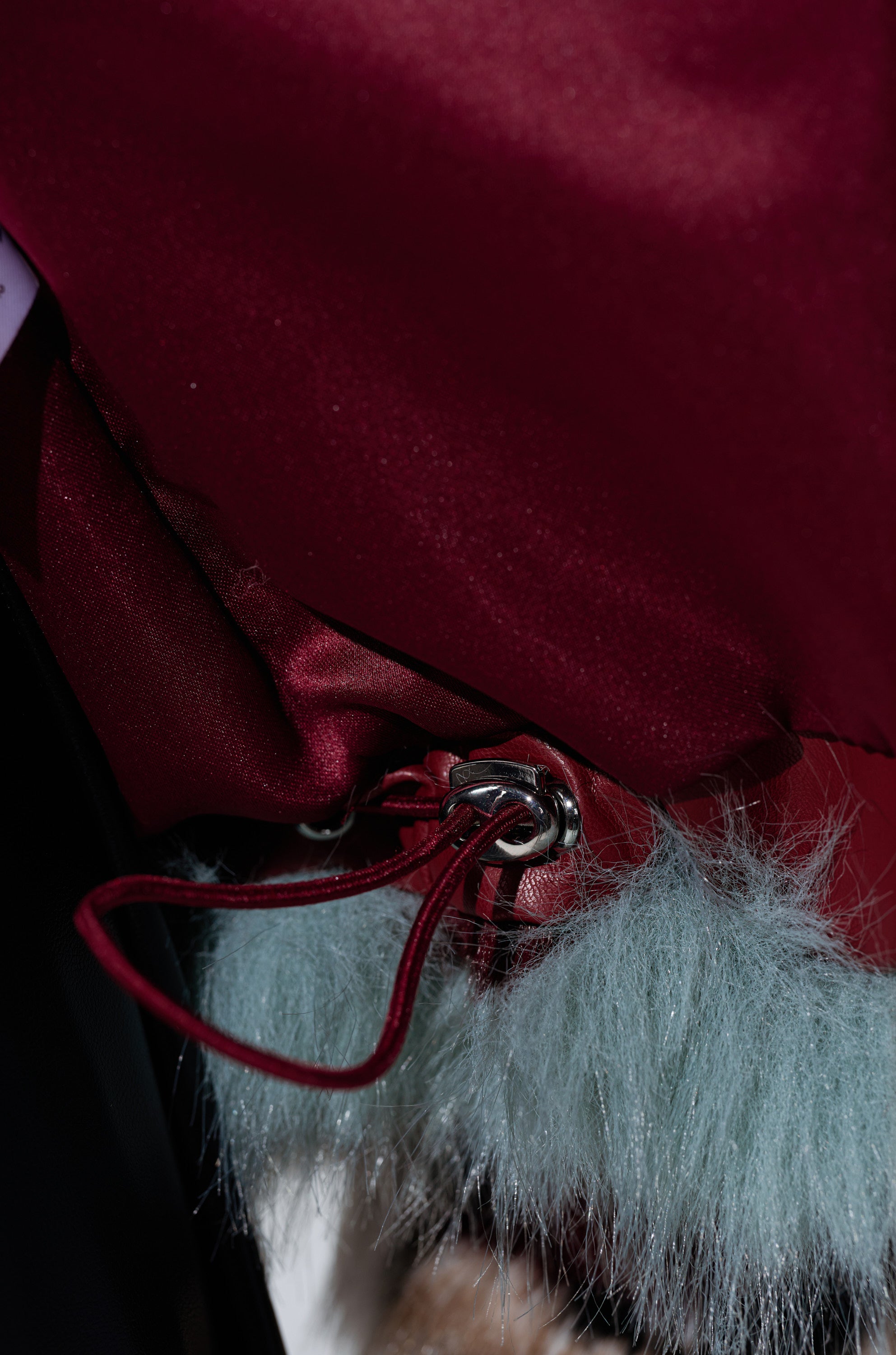 Close-up of a burgundy satin fabric with a silver clasp and red leather strap, attached to light blue-gray faux fur from the HAVE IT ALL PANELED FUR COAT.