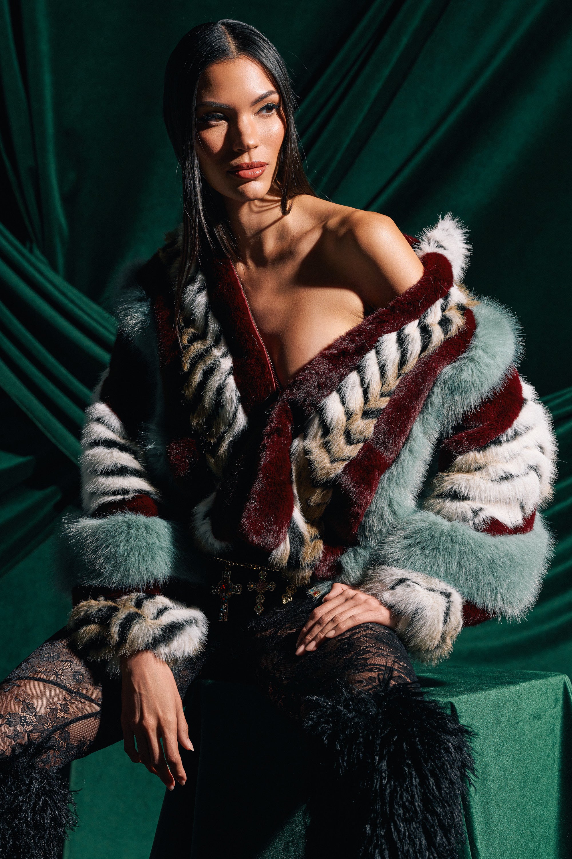 A woman with long dark hair poses against emerald green drapes, wearing the HAVE IT ALL PANELED FUR COAT in maroon, white, grey, and green panels, paired with lace-patterned black stockings.