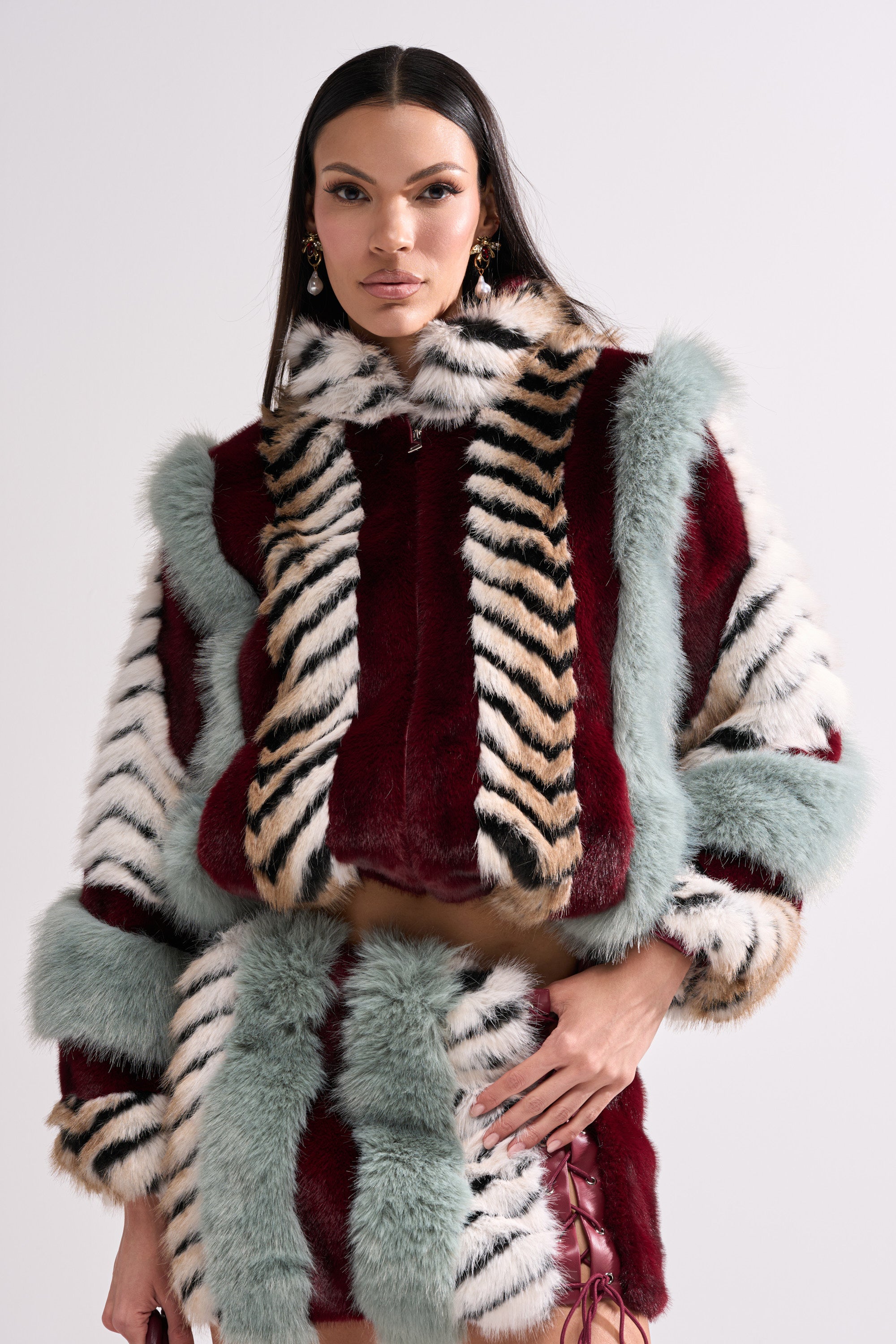 A woman with long dark hair and statement earrings showcases the HAVE IT ALL PANELED FUR COAT, featuring zebra stripes and panels of burgundy, tan, white, and mint green, as she poses confidently against a plain light background.