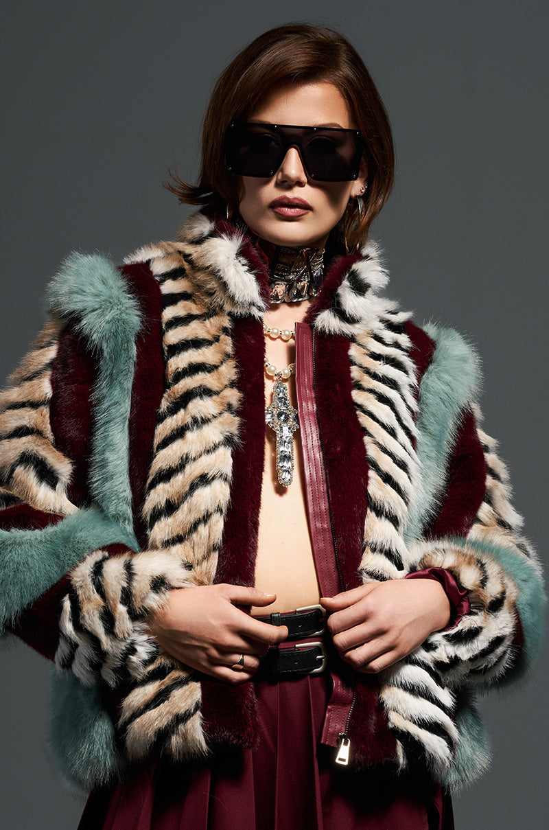 A person wearing oversized sunglasses poses confidently in the vibrant HAVE IT ALL PANELED FUR COAT adorned with bold patterns. They accessorize with a luxurious necklace and earrings, complementing their dark red outfit against a muted gray background.