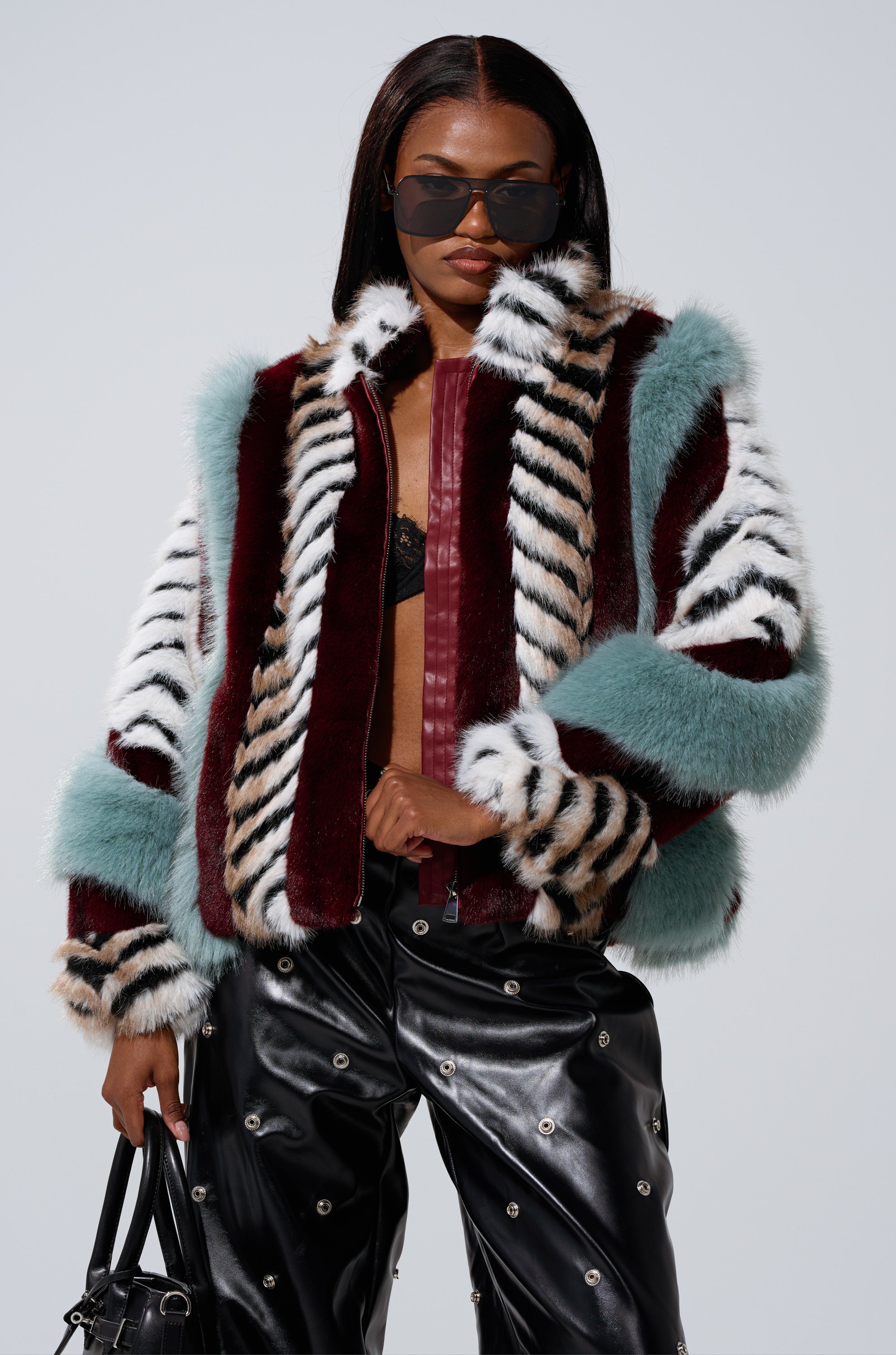 A woman in dark sunglasses wears the HAVE IT ALL PANELED FUR COAT—multicolored with teal, burgundy, white, and striped panels—paired with black leather pants featuring silver eyelets and a small black handbag.