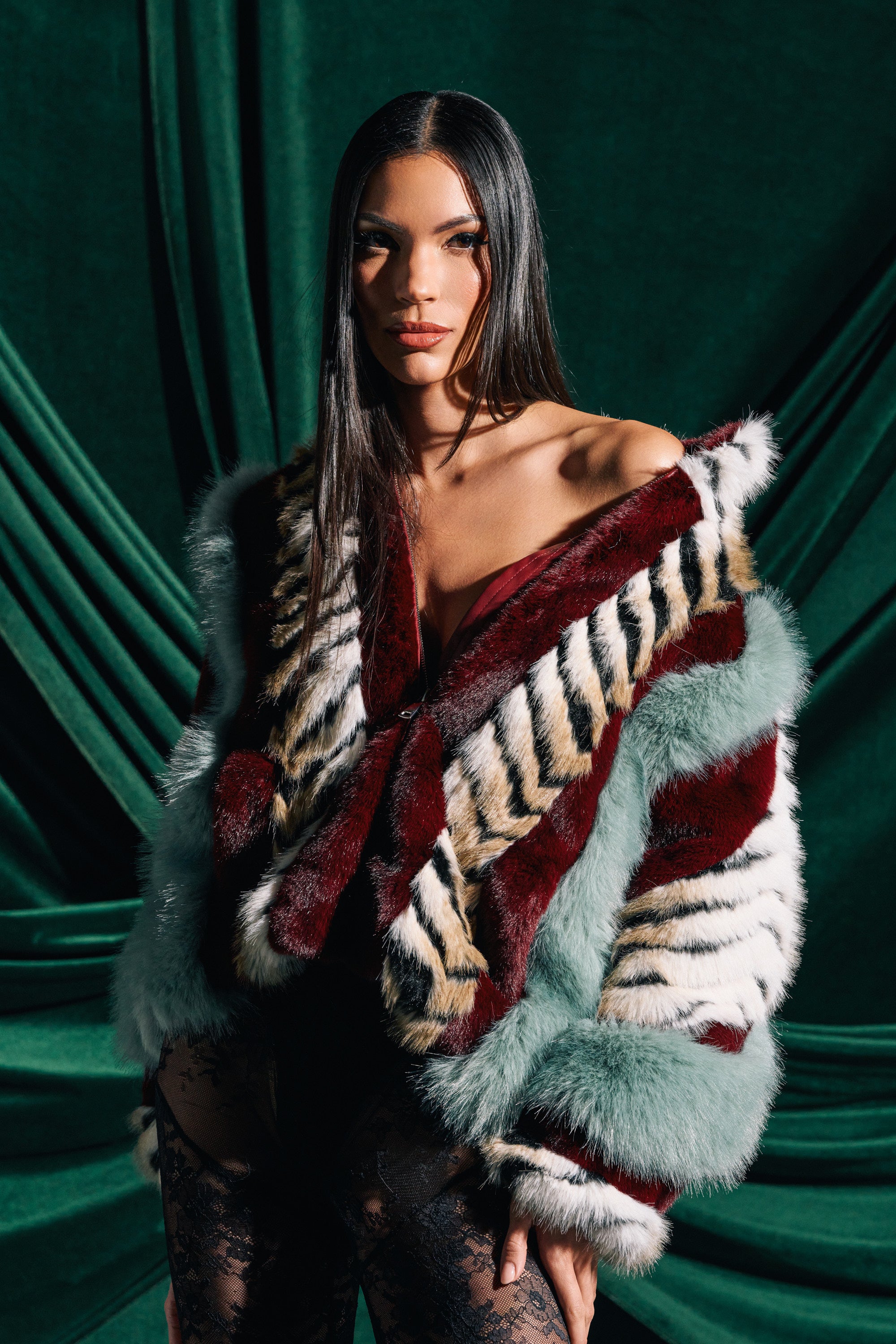 A woman with long straight hair poses before dark green drapes, wearing the HAVE IT ALL PANELED FUR COAT featuring bold red, white, black, and pale blue stripes over a black lace outfit.