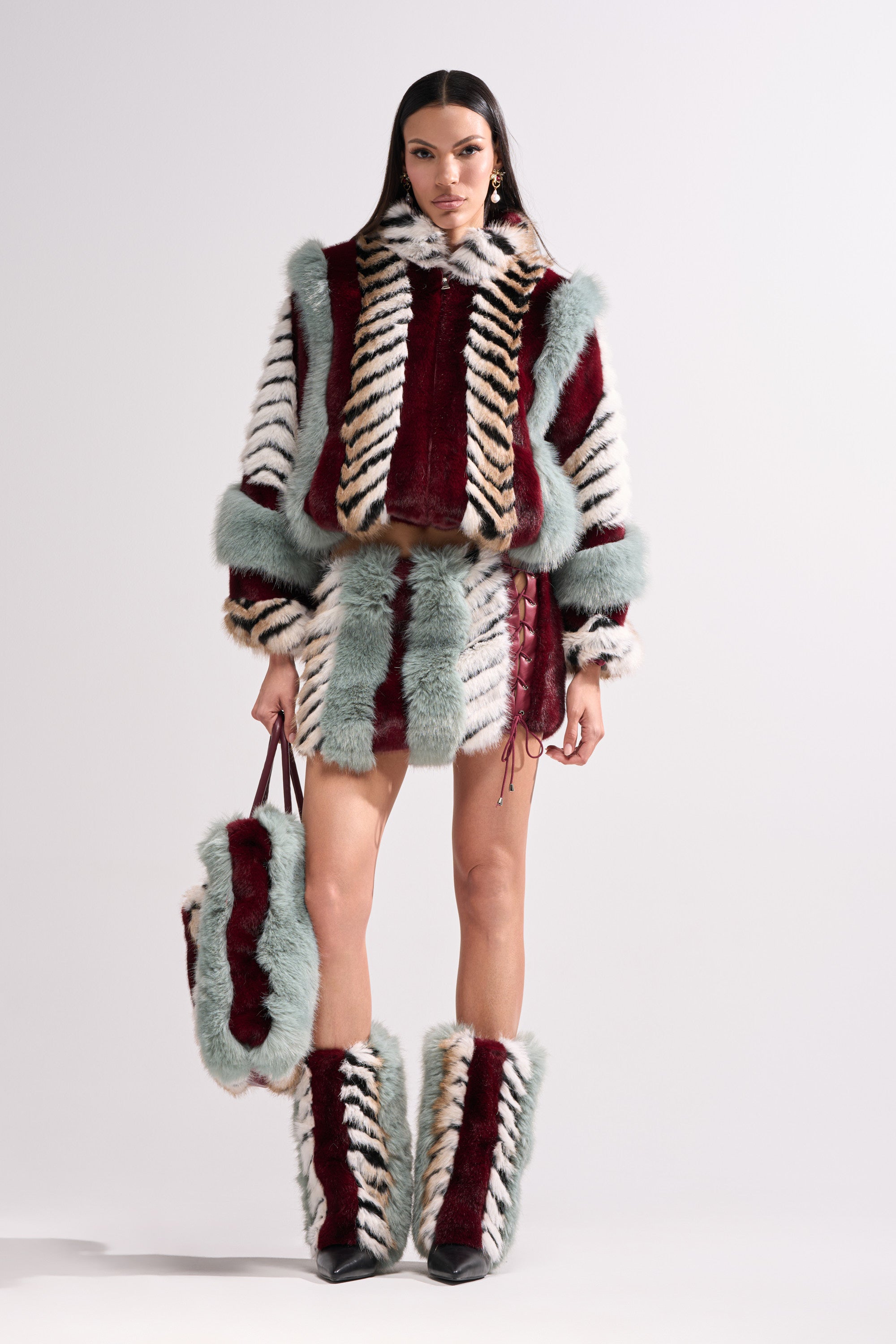 A woman poses against a plain light background in the HAVE IT ALL PANELED FUR COAT, a bold cropped faux fur jacket with burgundy, white, black, and mint green color blocks, styled with a mini skirt, tall boots, and a matching handbag.