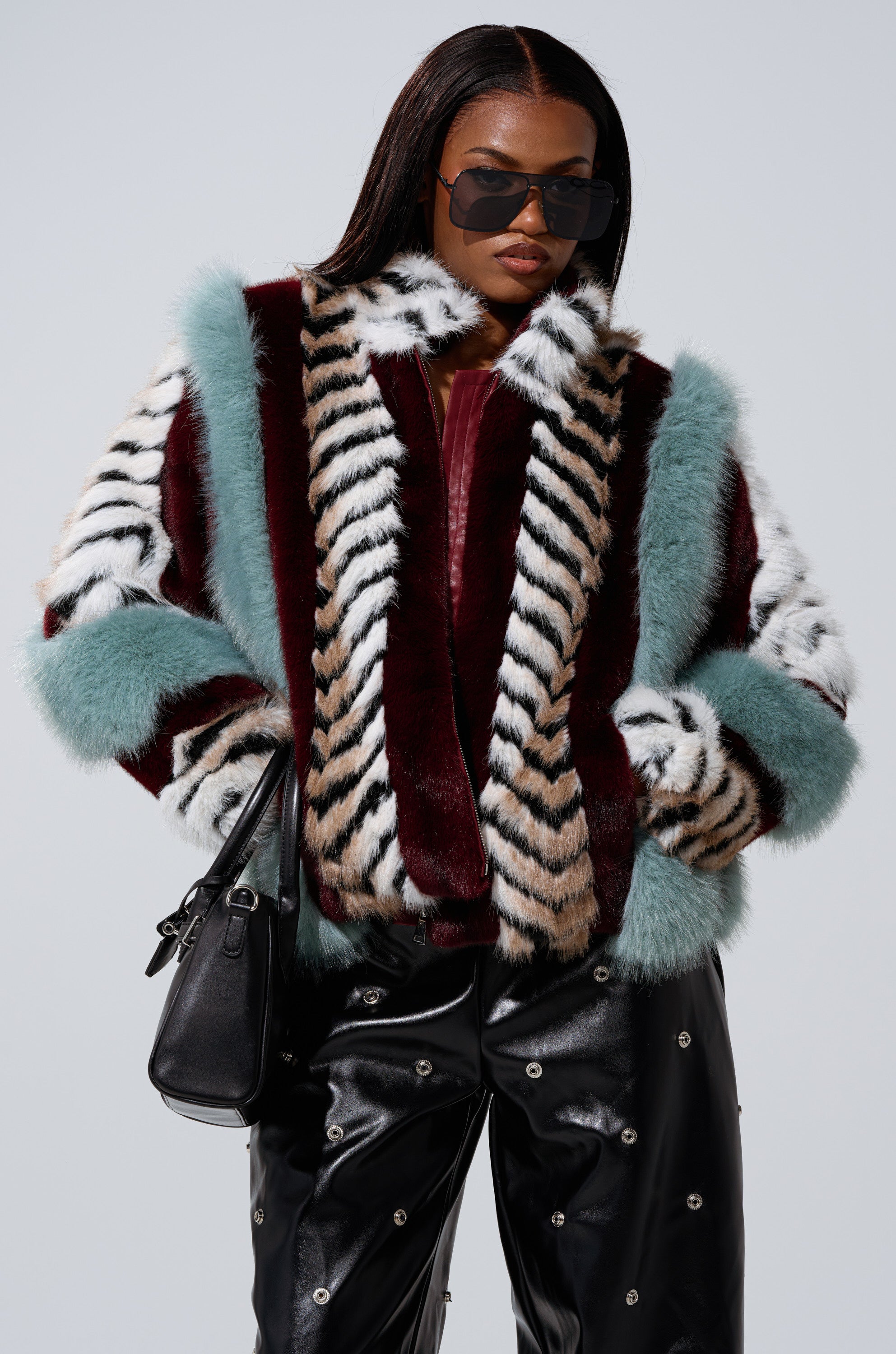 A woman wears the HAVE IT ALL PANELED FUR COAT featuring teal, burgundy, and striped panels, paired with black leather pants with silver accents, black sunglasses, and a black handbag.