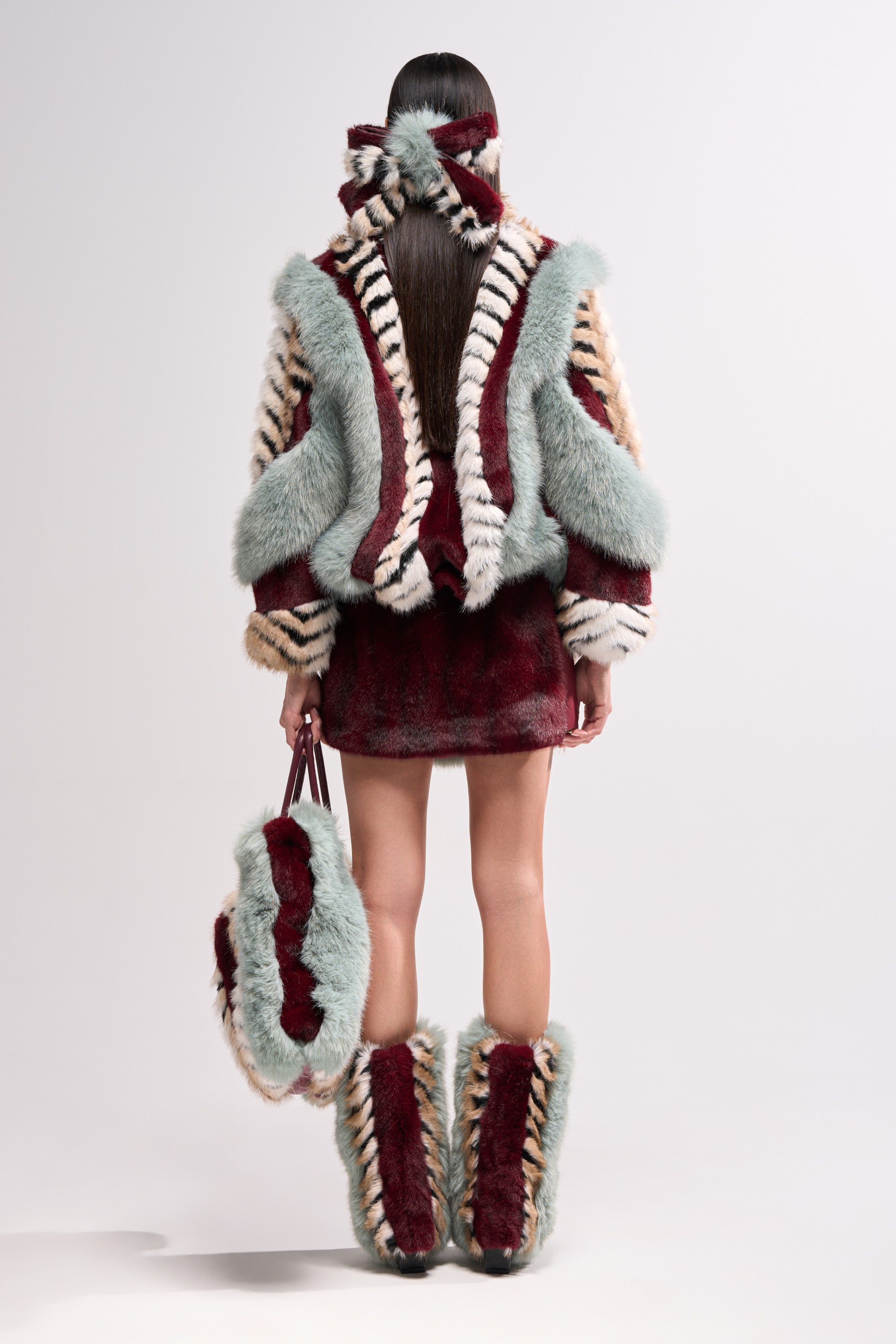 A person with long straight hair, seen from behind, wears the HAVE IT ALL PANELED FUR COAT in burgundy, pale blue, and cream with a short skirt, knee-high boots, and a matching handbag for a bold textured look.