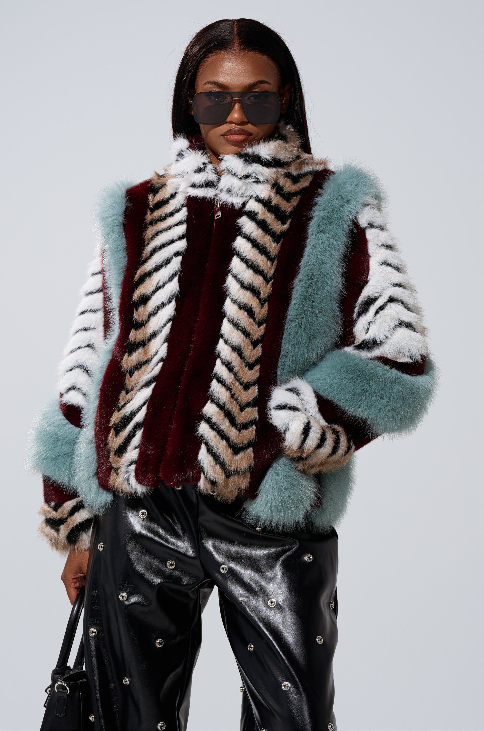 A person wearing large black sunglasses, black leather pants with silver embellishments, a black handbag, and the HAVE IT ALL PANELED FUR COAT in teal, burgundy, white, and beige stripes.