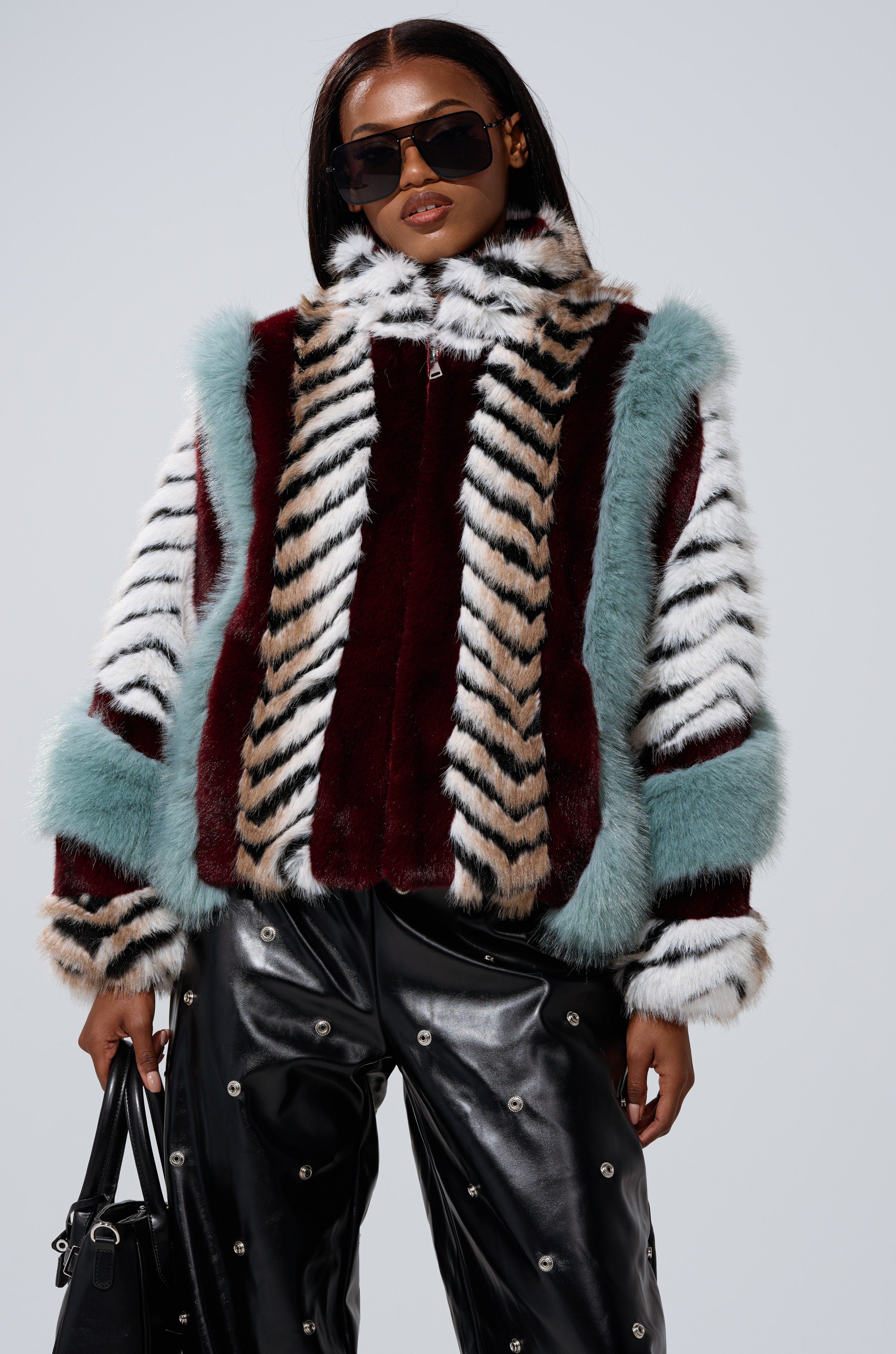 Wearing the HAVE IT ALL PANELED FUR COAT with burgundy, turquoise, and beige stripes, this person pairs it with studded black leather pants, a black bag, and oversized dark sunglasses.