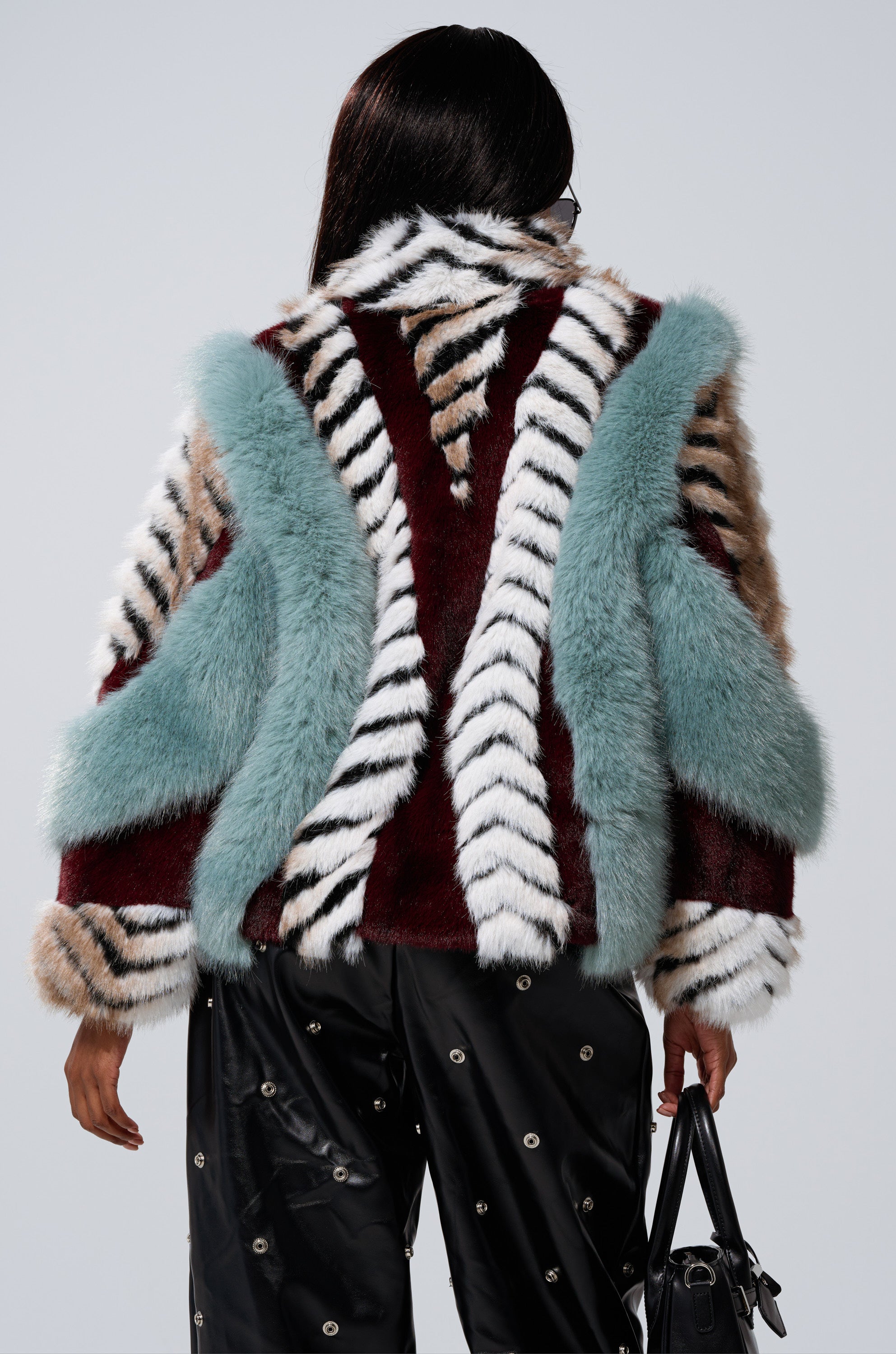 Seen from behind, a person with long dark hair wears the HAVE IT ALL PANELED FUR COAT—an eye-catching cropped fur jacket in teal, white, black, and brown—paired with black pants with metallic eyelets and carrying a black handbag.
