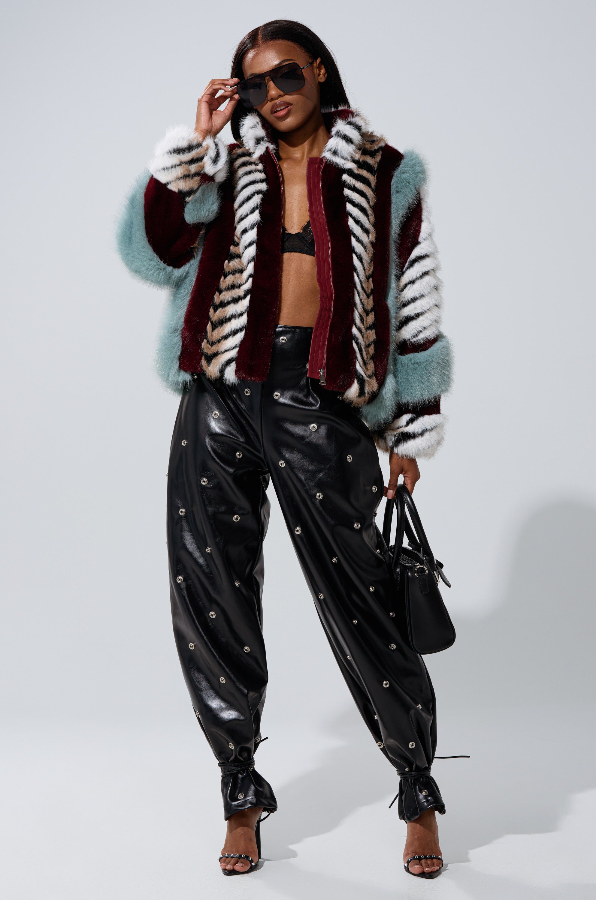 A woman wearing sunglasses, a black bralette, high-waisted leather pants with metal eyelets, heeled sandals, and holding a black handbag poses confidently in the vibrant HAVE IT ALL PANELED FUR COAT against a plain background.