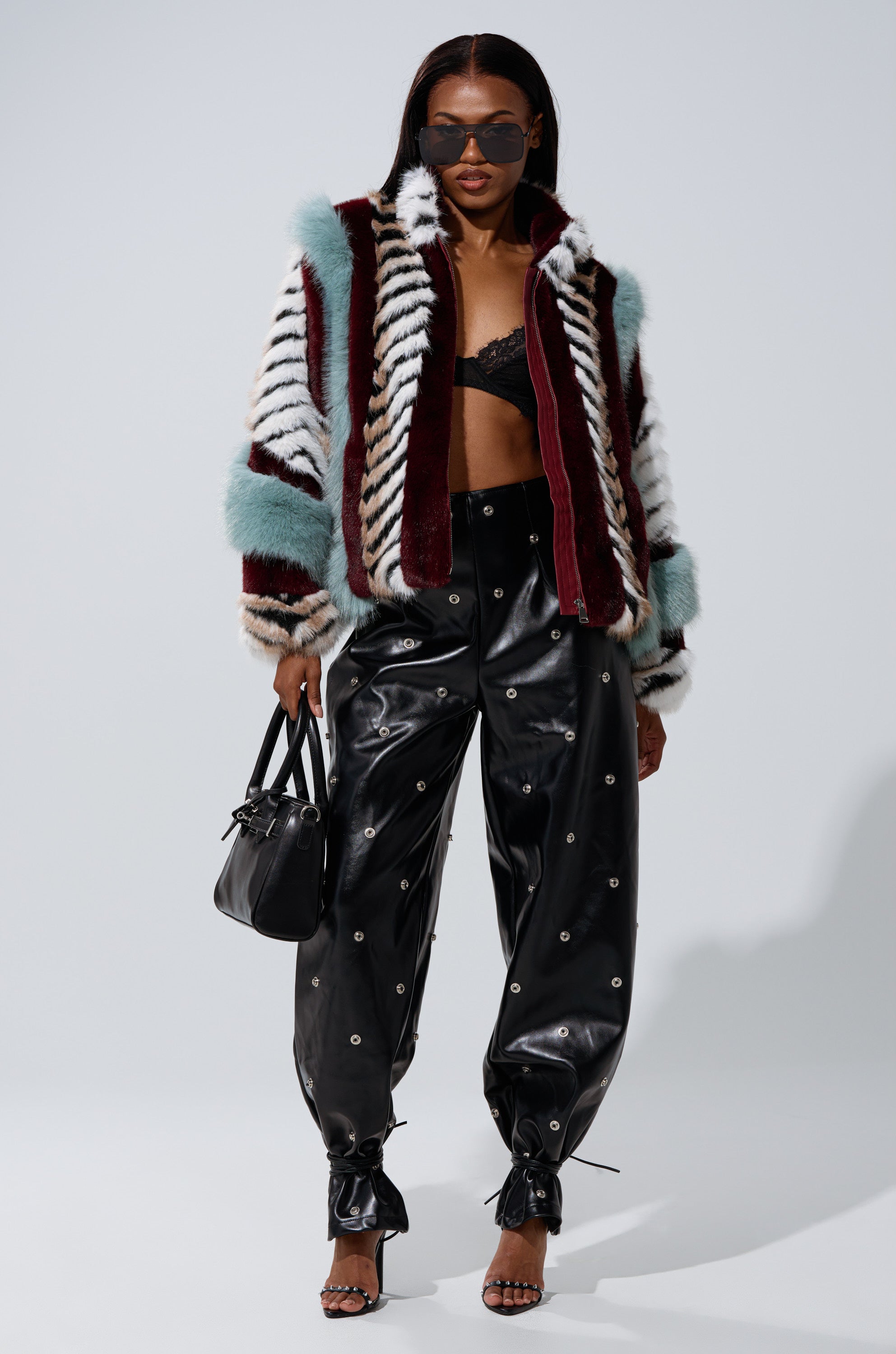 A woman wears the HAVE IT ALL PANELED FUR COAT in vibrant shades, sunglasses, a black lace bralette, high-waisted studded black leather pants, strappy heels, and carries a small black handbag against a plain backdrop.