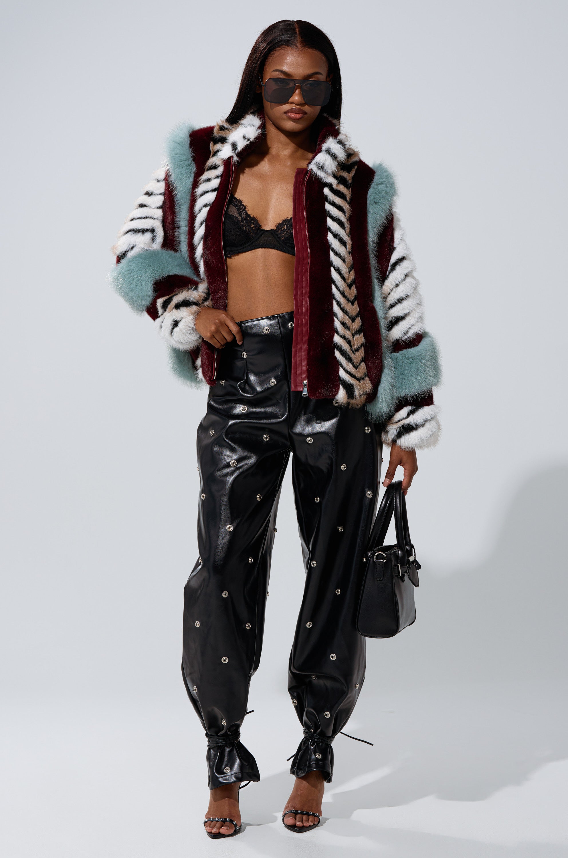A woman wears the HAVE IT ALL PANELED FUR COAT with maroon, teal, and white stripes, a black lace bralette, high-waisted black vinyl pants with silver grommets, black heeled sandals, sunglasses, and carries a sleek black handbag.