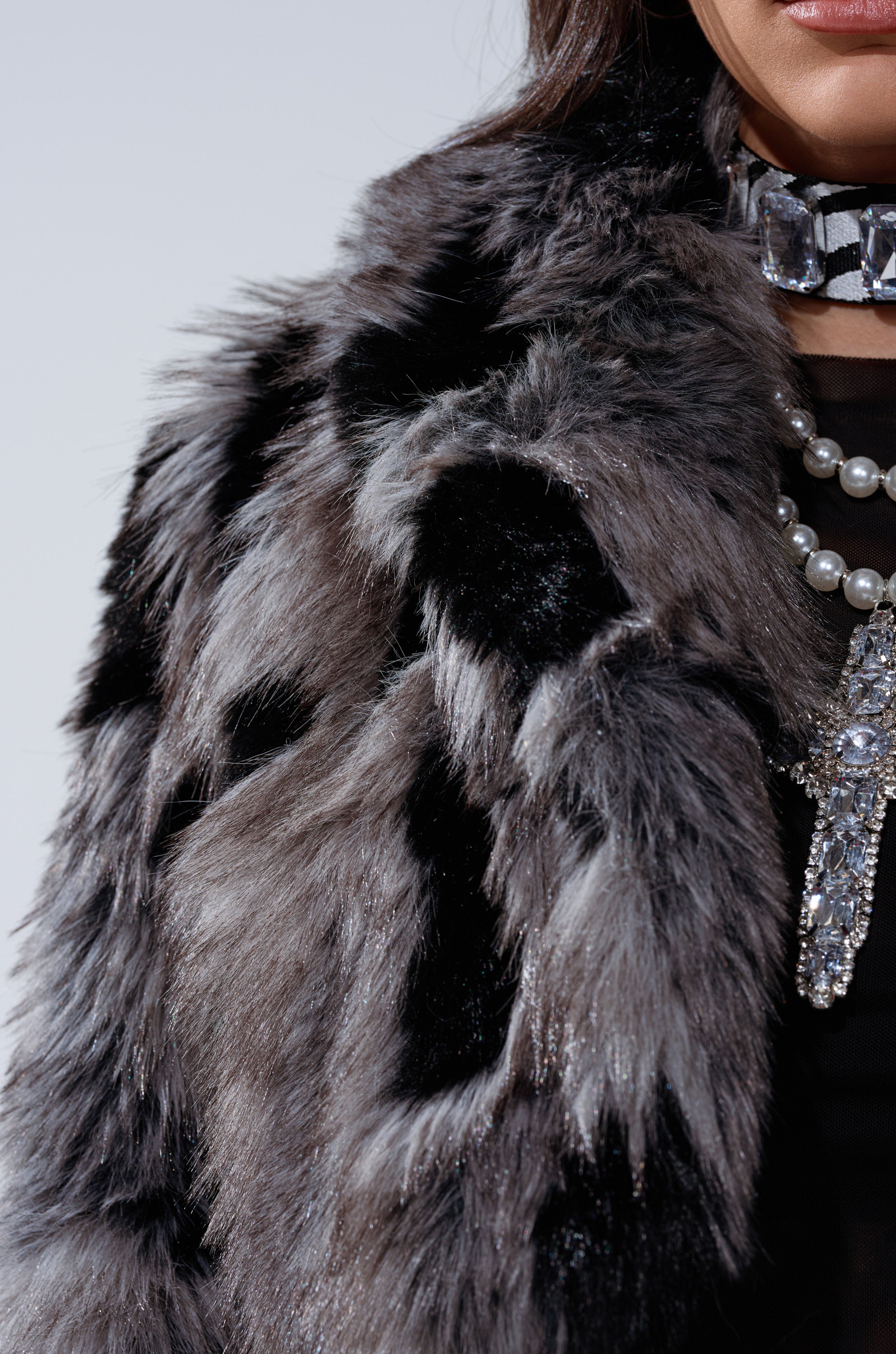 A person wears the AUSTIN CROPPED FAUX FUR IN GREY jacket in grey and black, styled with a jeweled choker and a pearl necklace with a sparkling pendant. Only their lower face and upper torso are shown.