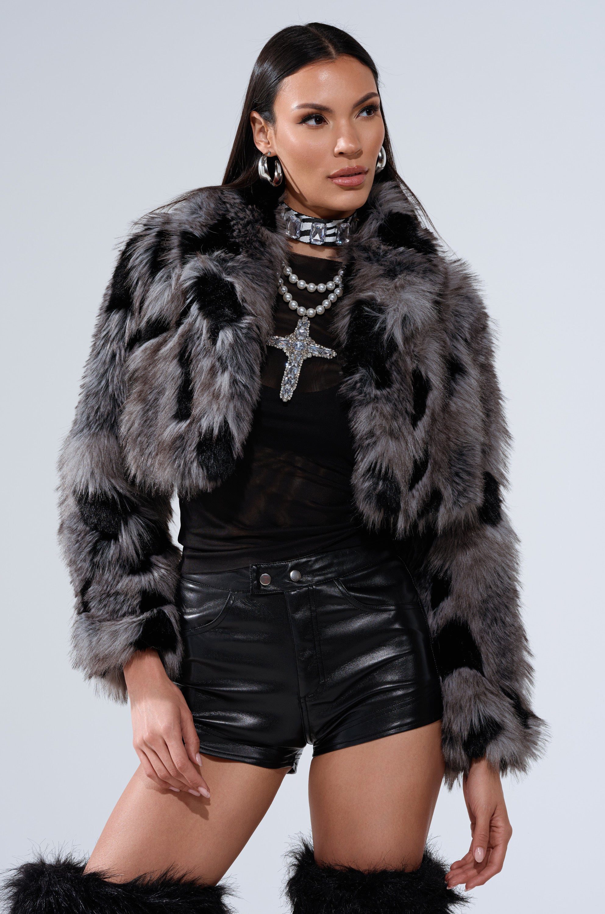 A woman wearing the AUSTIN CROPPED FAUX FUR IN GREY jacket, black leather shorts, a sheer black top, silver layered necklaces, hoop earrings, and matching furry boots poses confidently against a plain light background.