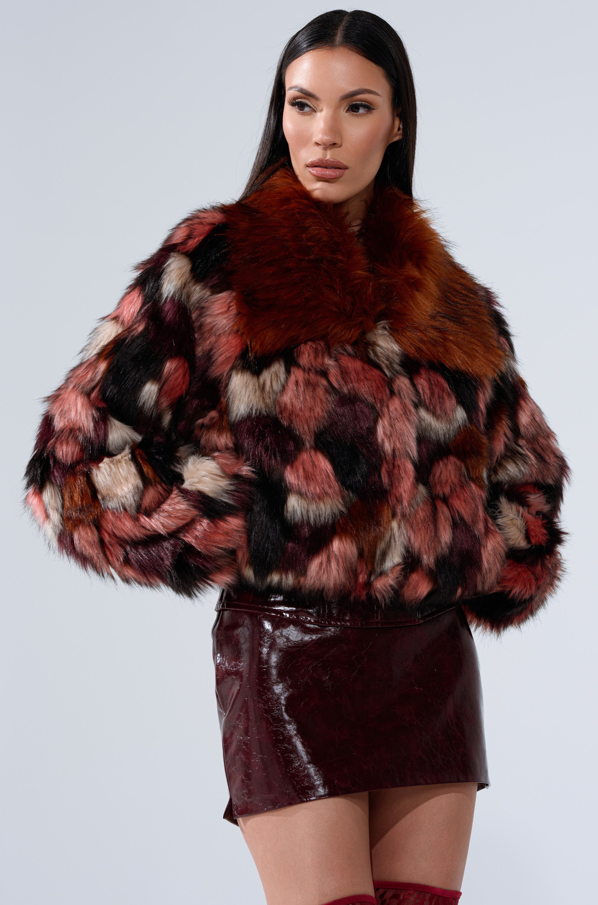 A woman models the DELTA FAUX FUR COAT, featuring red, black, and white patchwork, styled with a dark red shiny mini skirt against a plain light background.