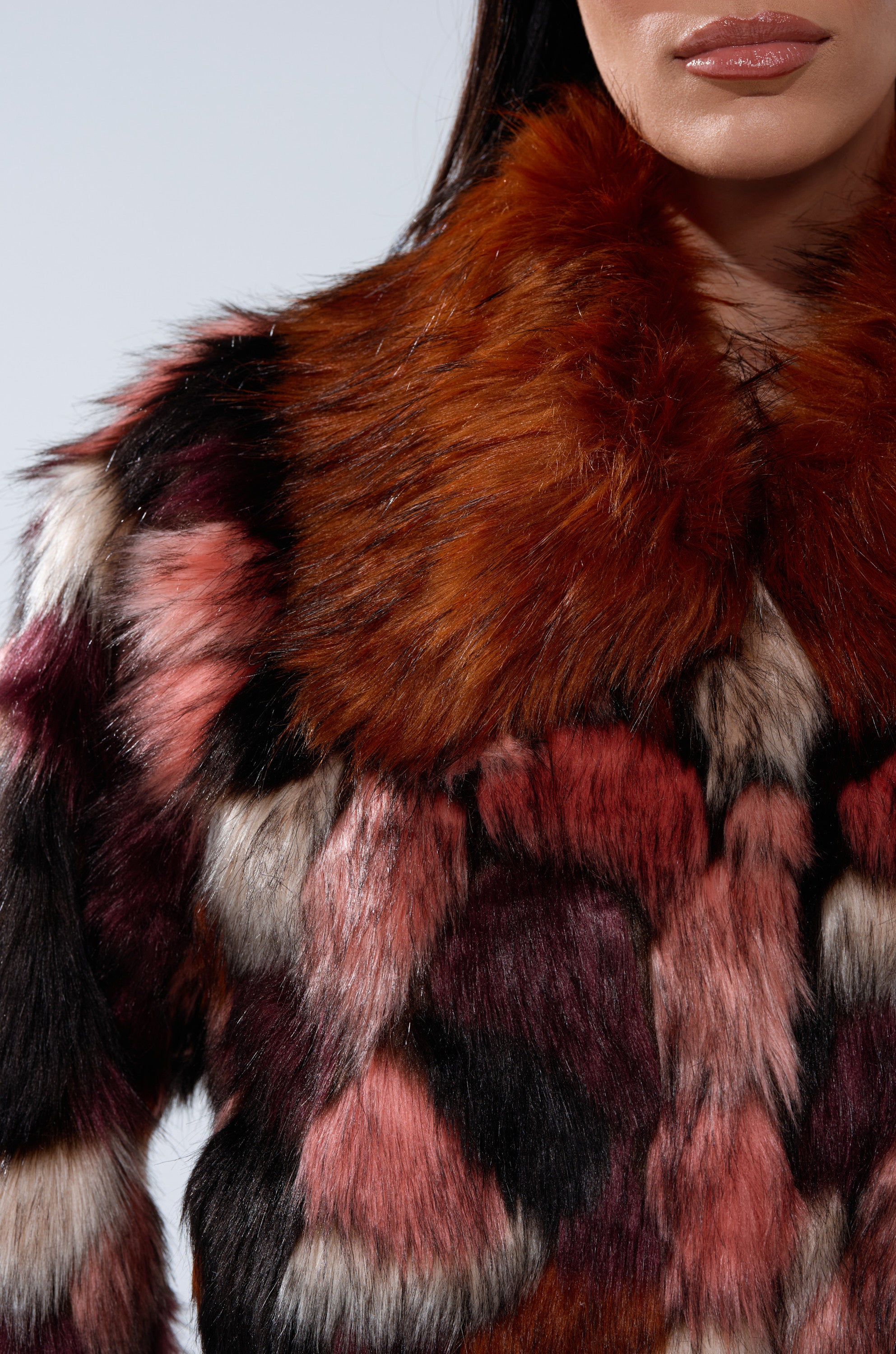 A woman wears the DELTA FAUX FUR COAT, a cropped design featuring a vibrant patchwork of brown, red, black, and white with a large fluffy collar. Only her lower face and upper torso are shown against a plain background.