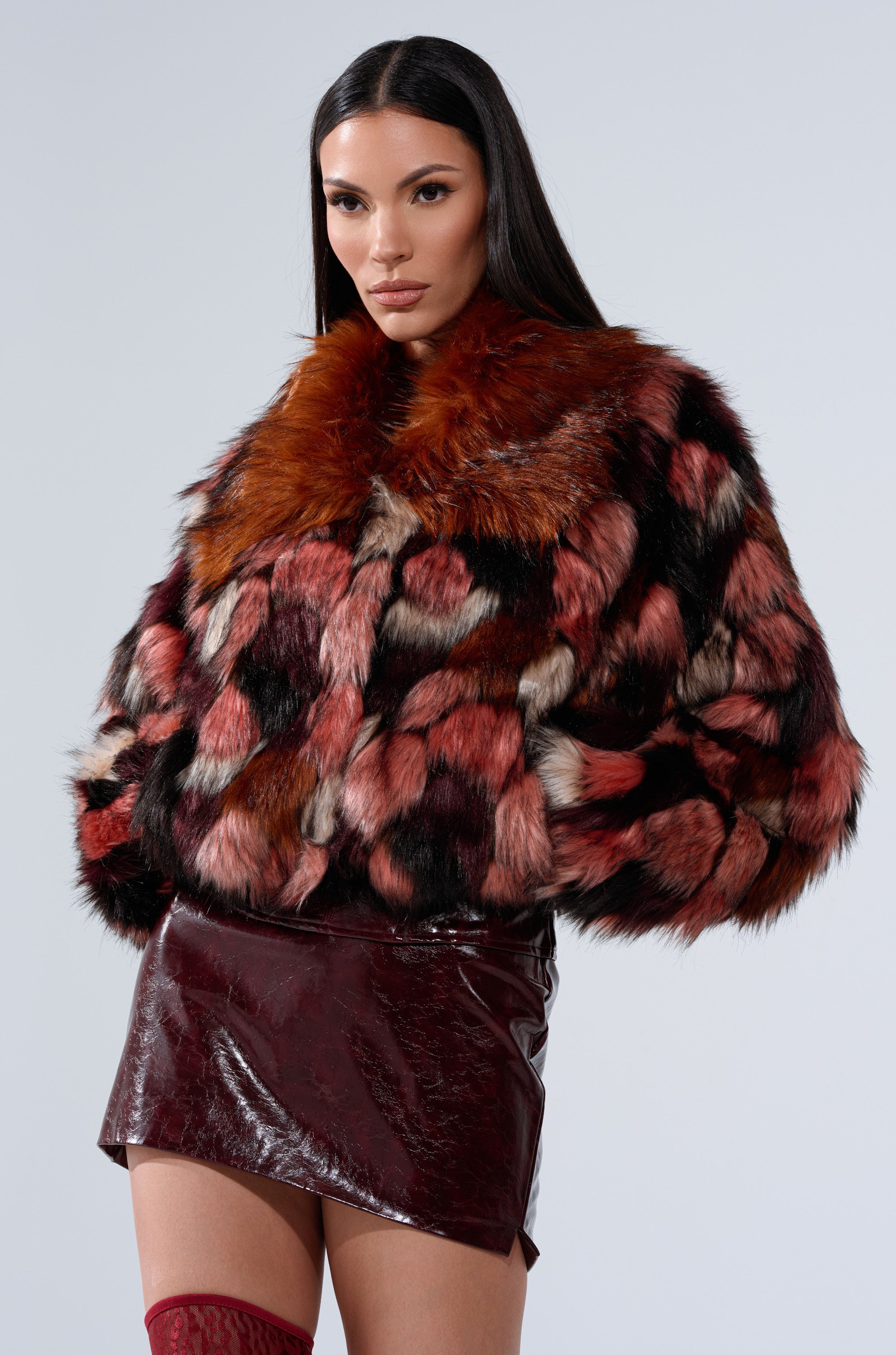 A woman with long dark hair wears the DELTA FAUX FUR COAT in red, black, and brown patchwork over a shiny maroon mini skirt and red stockings, standing against a plain light background.