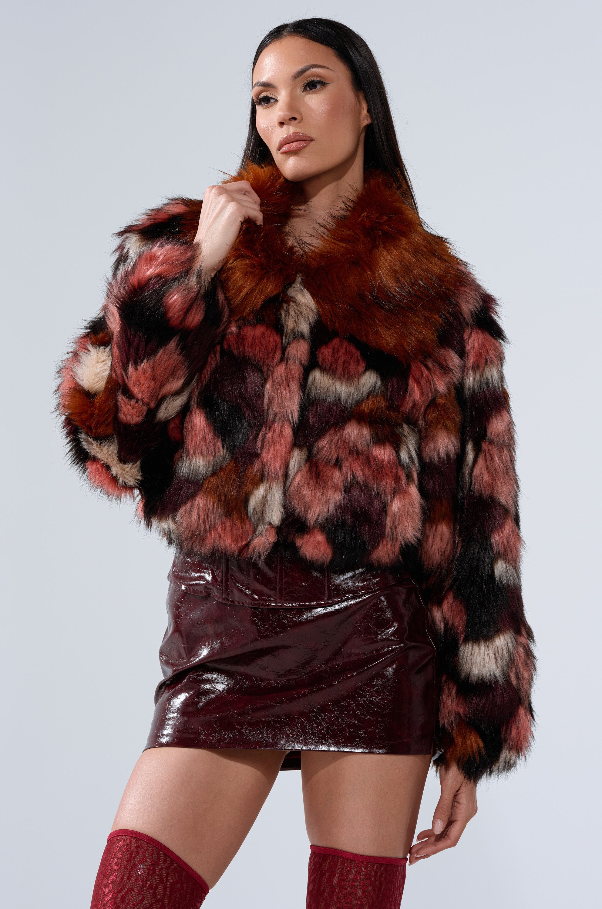 A woman stands confidently against a light gray background, wearing the DELTA FAUX FUR COAT with red, black, and white patches, paired with a shiny burgundy mini skirt and red knee-high socks.