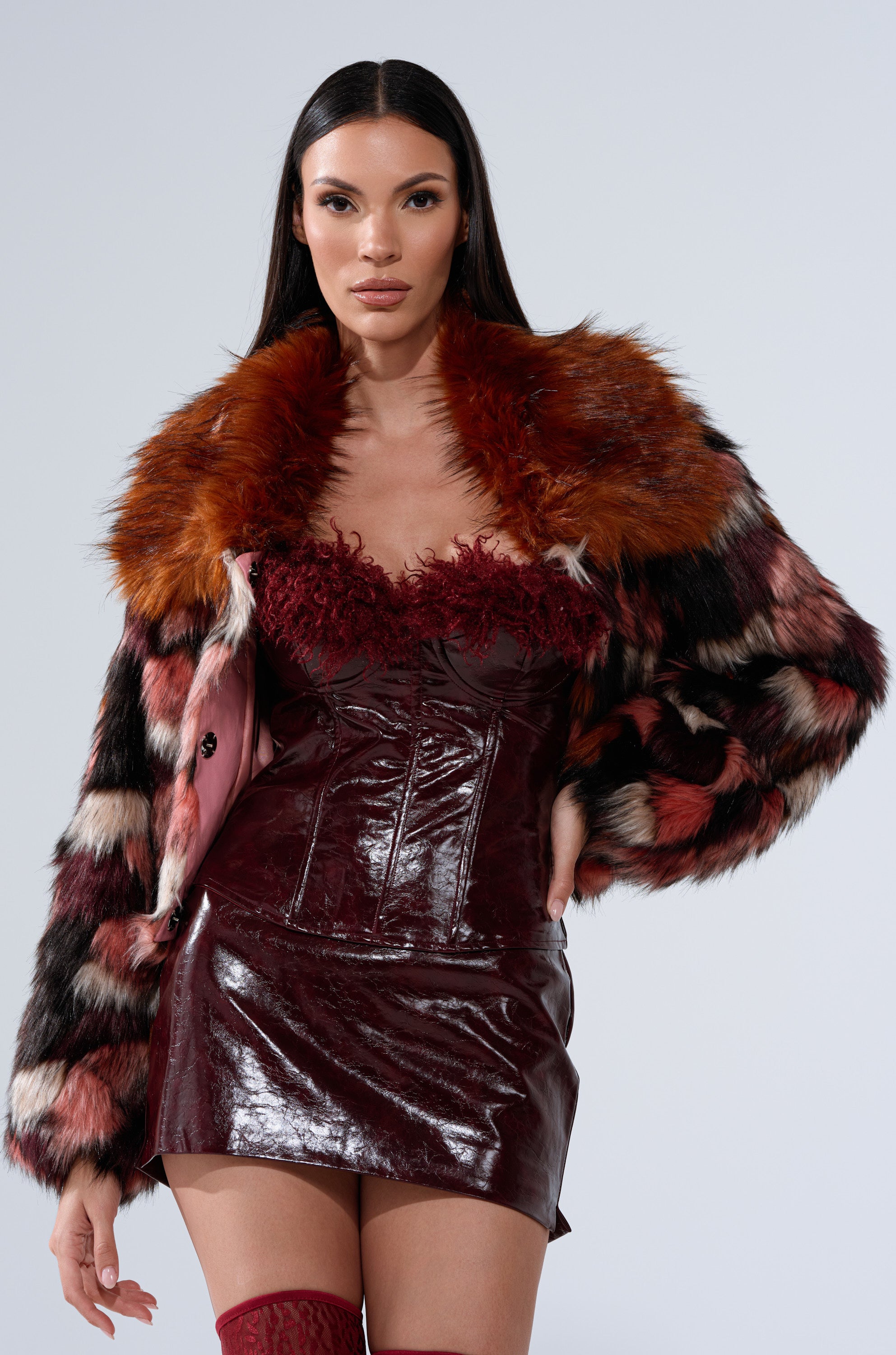 A woman with long dark hair models the DELTA FAUX FUR COAT in black, red, and pink over a shiny burgundy corset dress, standing confidently against a plain light background.