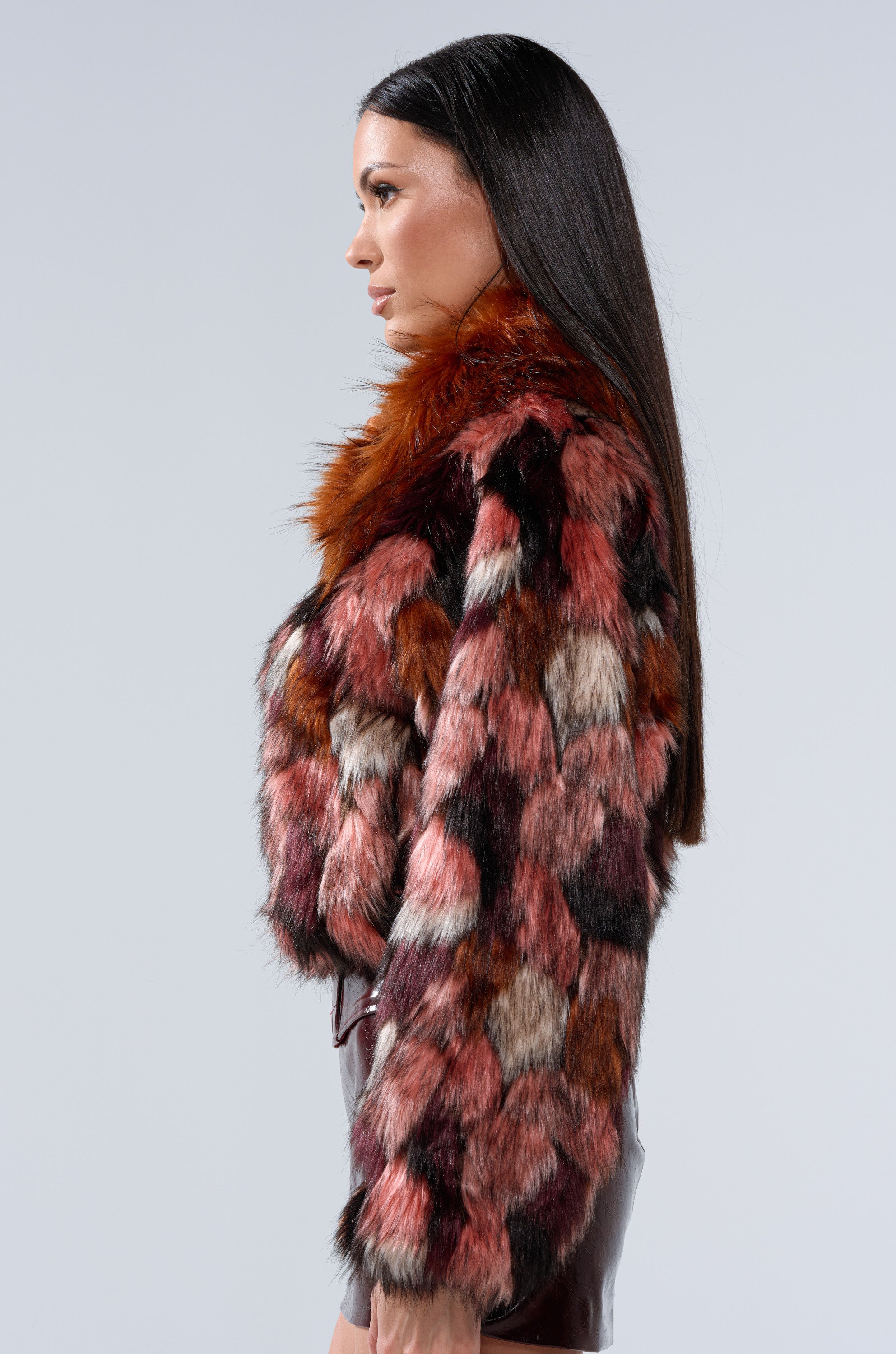 A woman with long straight dark hair, shown in profile, wears the DELTA FAUX FUR COAT, featuring a colorful patchwork design in red, pink, brown, and black shades over a shiny skirt against a plain light background.