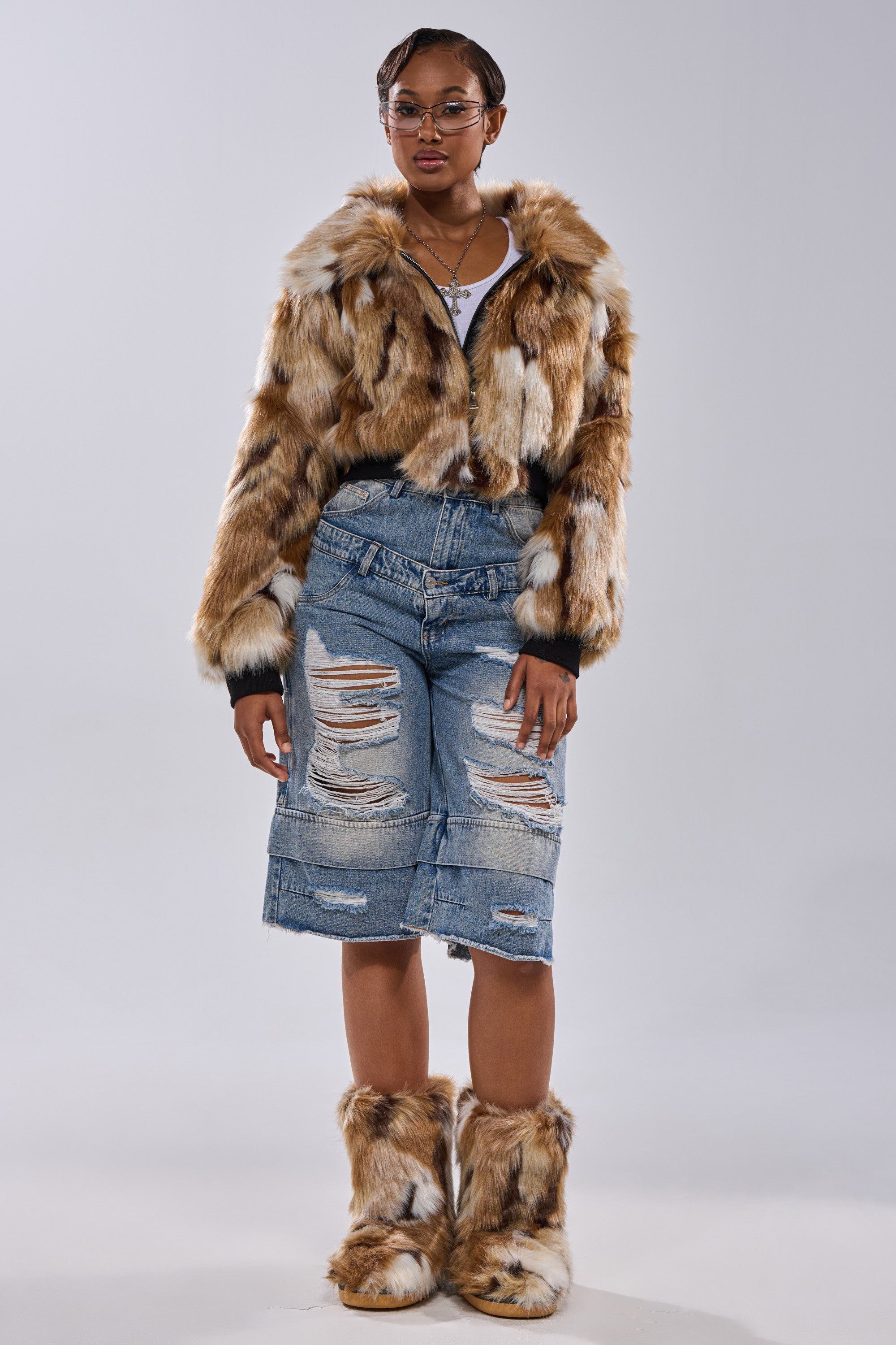 A person stands against a plain backdrop wearing the FENNEC FAUX FUR BOMBER in brown and white, matching fur boots, glasses, layered necklaces, and distressed denim knee-length shorts.