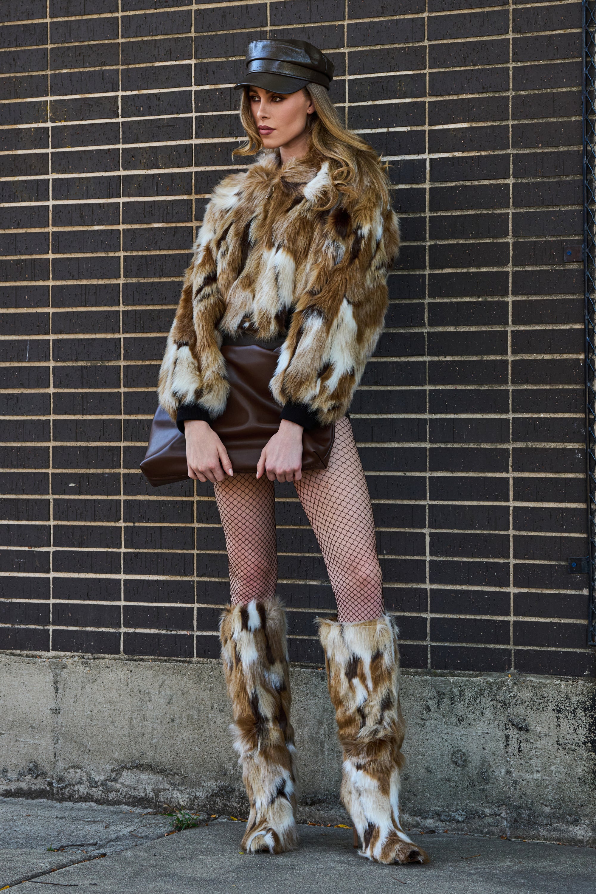 A woman stands against a brick wall wearing the FENNEC FAUX FUR BOMBER, matching fur boots, fishnet tights, a leather cap, and holding a large brown leather clutch. She looks to the side with confidence.
