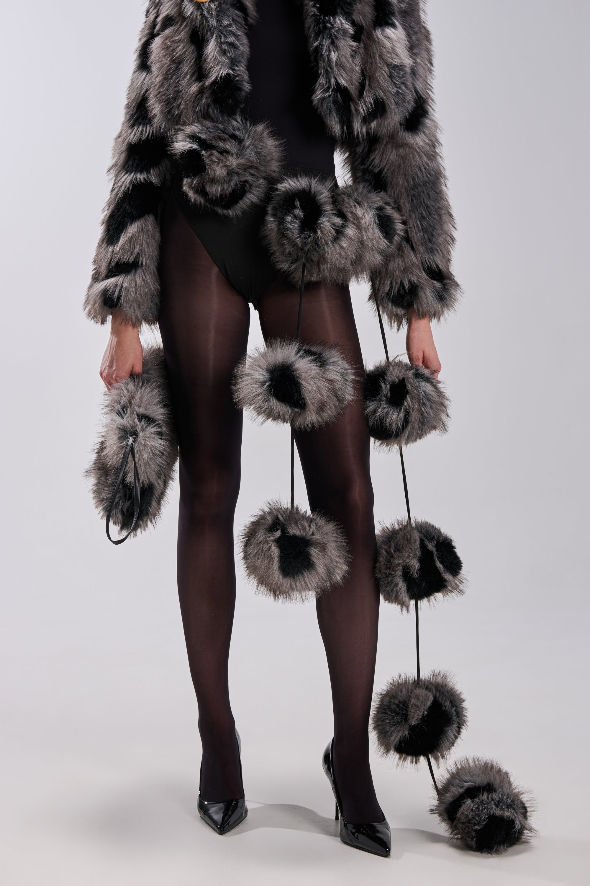 A person wearing a furry black and gray jacket, sheer black tights, and black heels stands with the BALLS TO THE WALL FAUX FUR SCARF—an oversized plush knit scarf with pom poms—draped around their body on long cords.