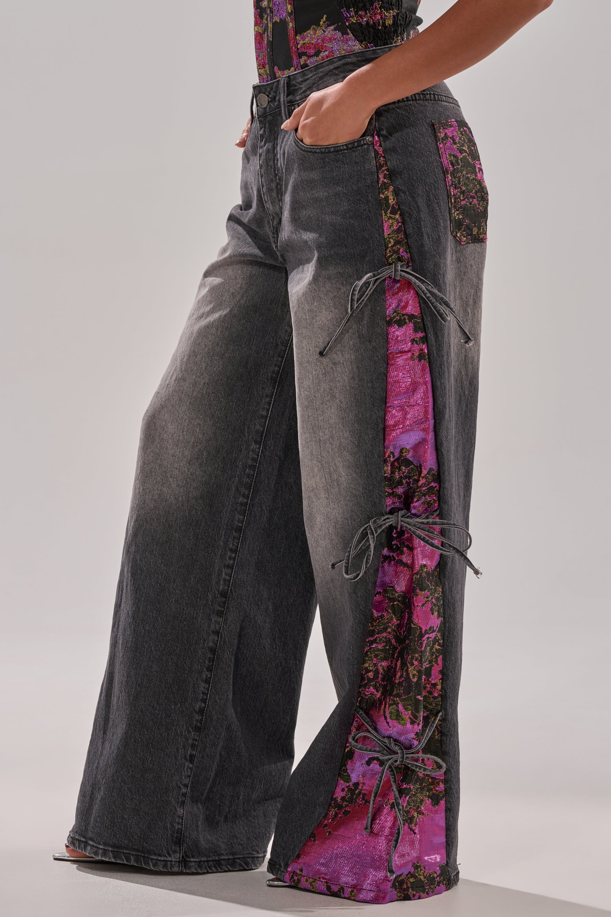 Someone wears LETS PARTY BROCADE RELAXED DENIM—wide-leg, dark gray jeans featuring vibrant pink and green floral side panels, ruched accents, metallic brocade details, and front pockets.